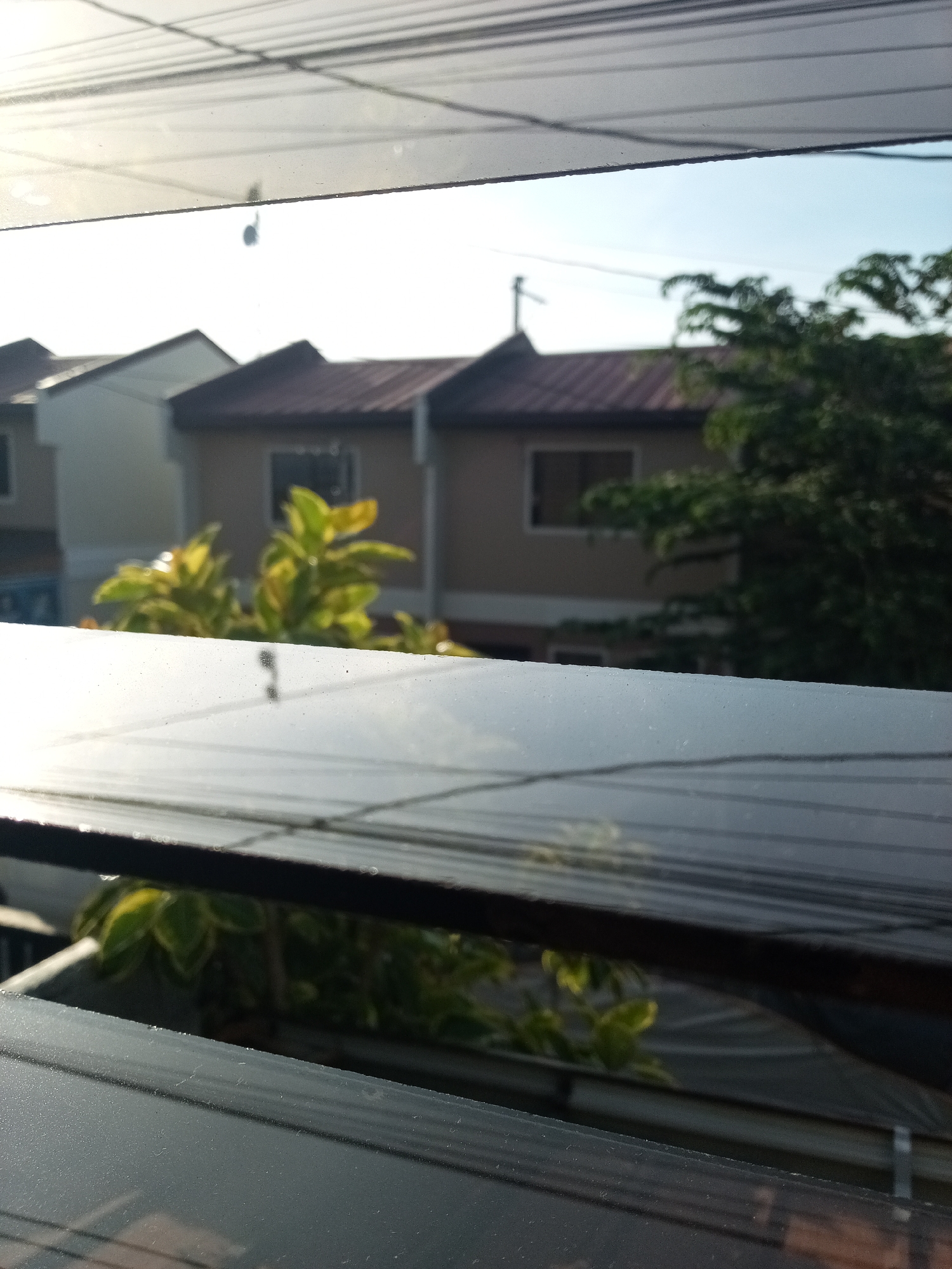 A slightly blurred view from inside a building in Davao City, Philippines, looking out through a glass window with horizontal slats. The window frame is visible. Outside, there are several houses, one painted a light beige with a red-brown roof, and another pale yellow. Green trees are visible, particularly to the right of the frame. An overhead wire is stretched across the sky. The sky is pale blue.