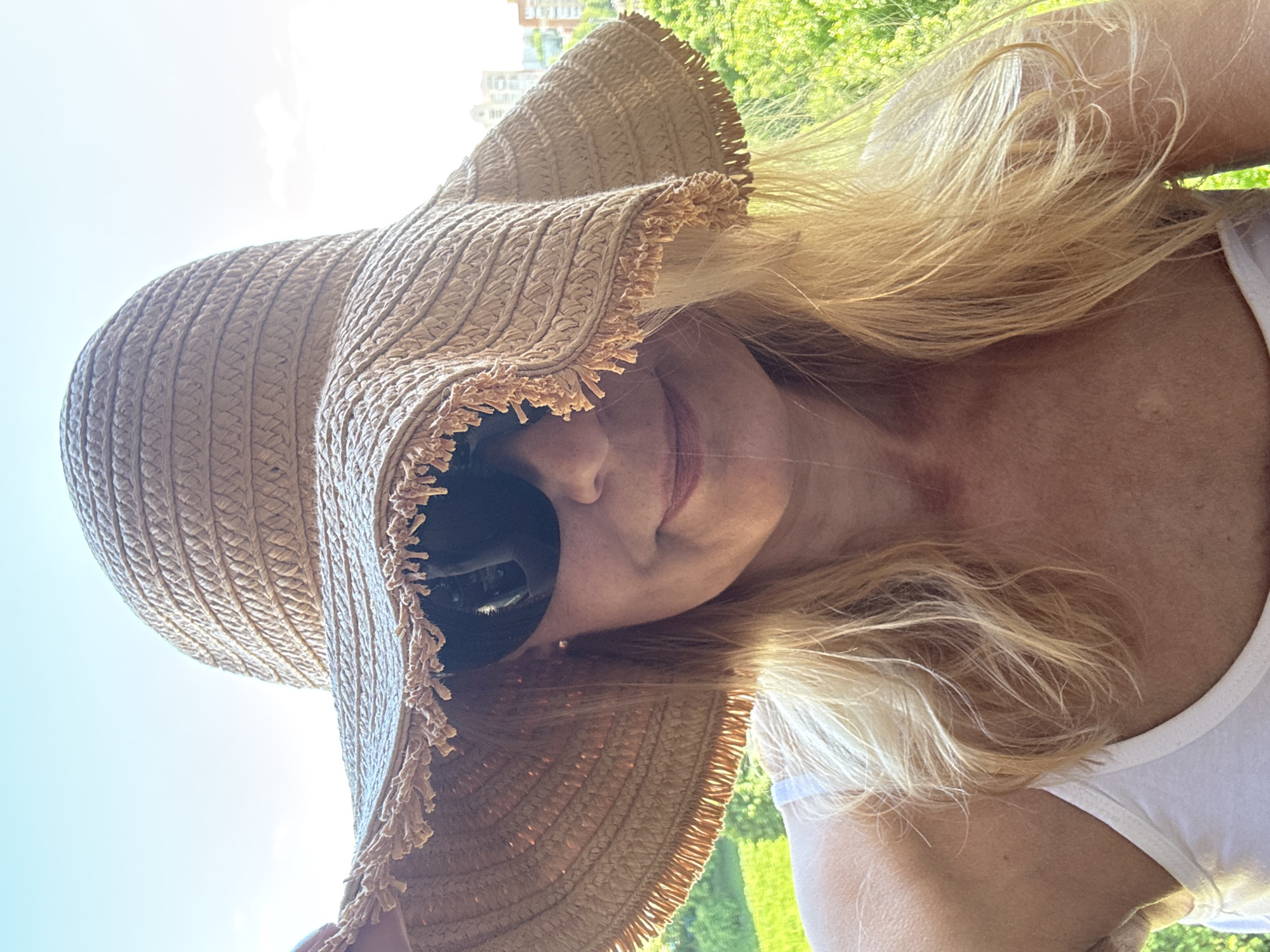 A woman with blonde hair is wearing sunglasses and a large straw hat in an outdoor setting. She is wearing a white tank top. Greenery is visible in the background. The location is Minsk, Belarus.