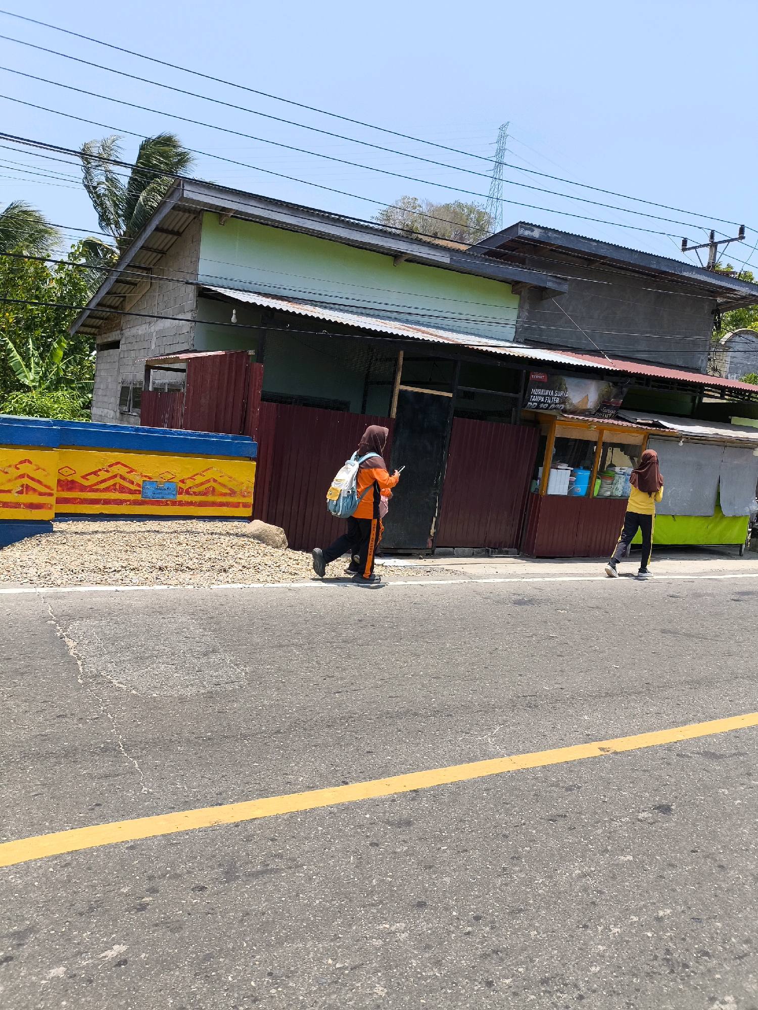 The image shows a street scene in front of a building with a corrugated metal roof. Two young women wearing hijabs and athletic clothing are walking on the sidewalk. The woman on the left is wearing an orange and black outfit with a backpack, and the woman on the right is wearing a yellow and black outfit. The building appears to be a small business, with signage that is partially visible. The street is paved with asphalt and has a yellow line marking the lane. The sky is clear and blue, and there are some trees in the background. A power line tower is visible in the distance. The scene appears to be in a tropical or subtropical climate, as indicated by the lush vegetation and the presence of palm trees. The overall impression is of a typical day in a small town or village in Indonesia.