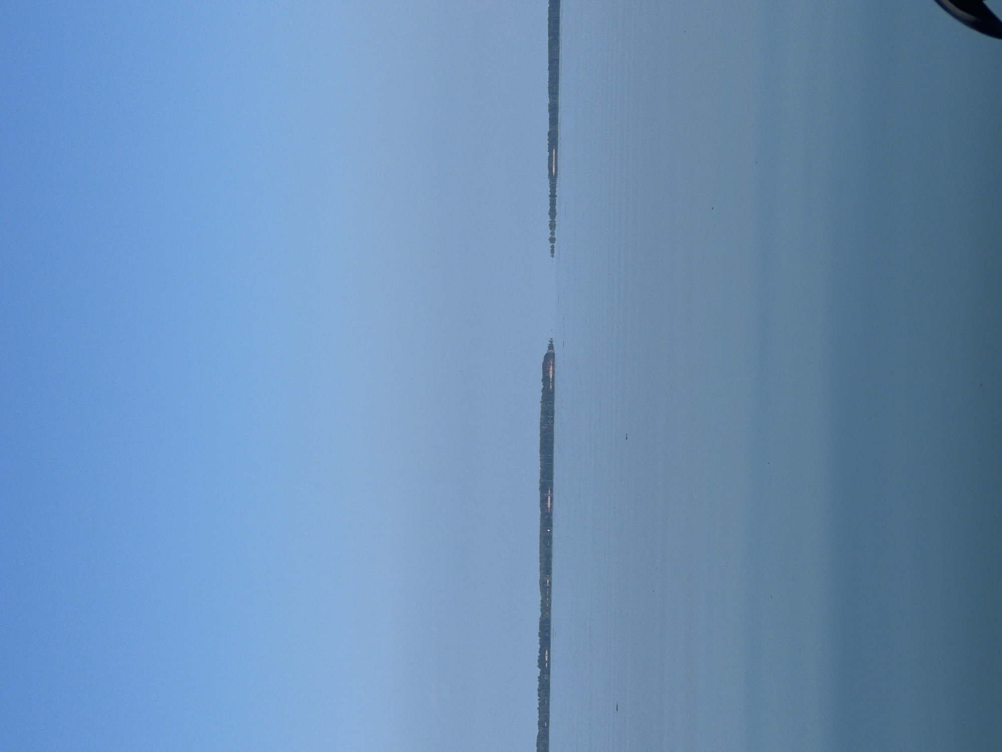 The image depicts a wide expanse of calm, light blue water under a clear, pale blue sky. The horizon is visible, with a thin line of green trees and some small structures visible on distant landmasses. There are two distinct landmasses in the distance, separated by a gap of water. The water surface is smooth, reflecting the sky. The overall scene conveys a sense of openness and tranquility. The location is identified as Beregovoe, Russia.