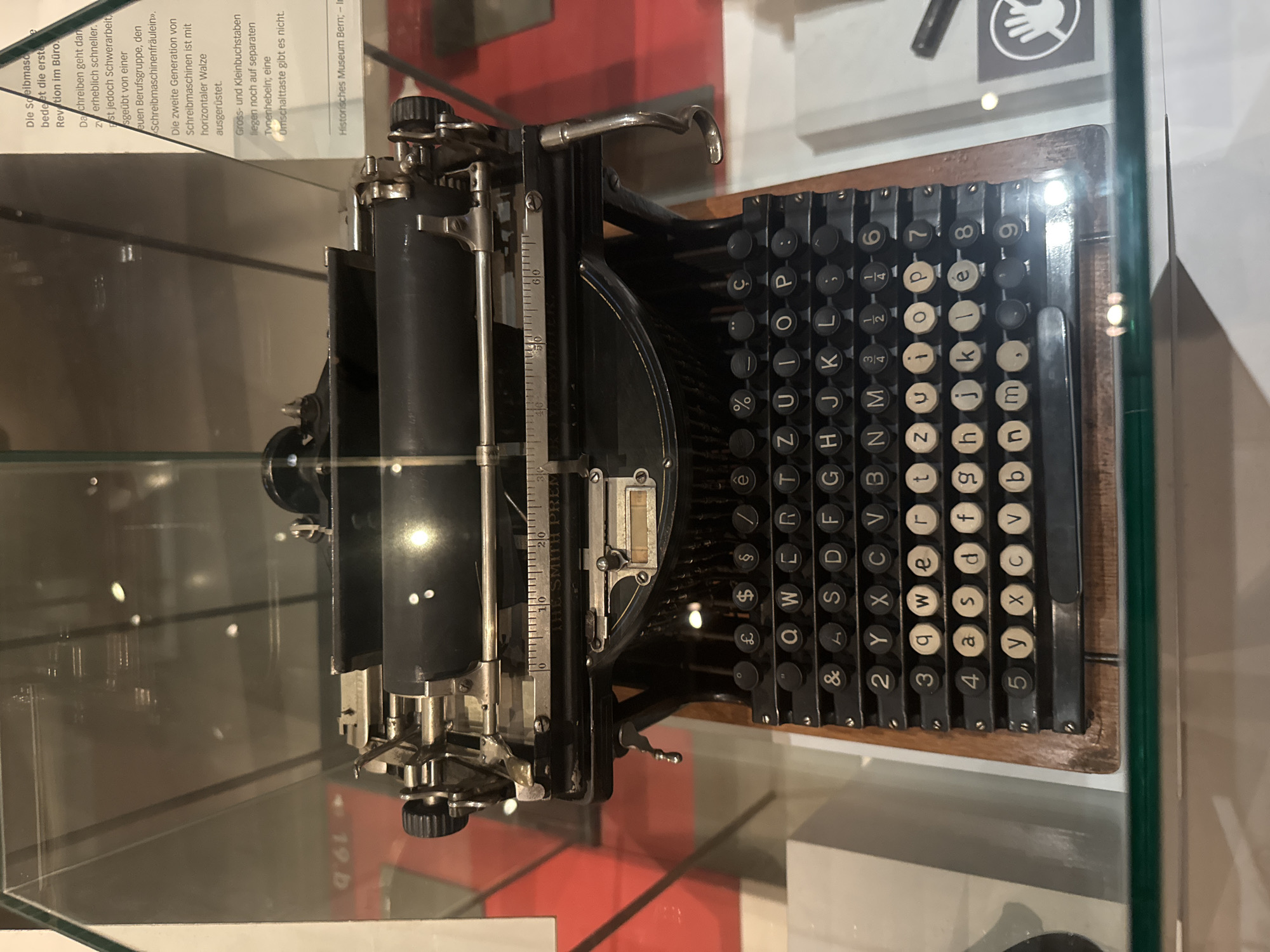 This media file shows a close-up, side-view of a vintage typewriter, likely displayed in a museum. The typewriter is black with silver metal accents and a wooden base. It features a full keyboard with circular keys, each bearing a letter, number, or symbol. Some keys are white, while others are black. The machine has a carriage with a roller and a ruler marking on its left side. Above the keyboard, a metal lever is visible.
In the background, through a glass enclosure, there are glimpses of exhibition displays. Text is visible on a white sign in the upper left, partially obscured by the typewriter. The text is in German and appears to be descriptive of the typewriter, mentioning "Speibmasc", "Revation im Büro", "Schreibmaschinen", and "Historisches Museum Bern".
The lighting suggests an indoor setting, possibly a museum exhibition, with bright reflections on the glass. No people are visible in the frame. The overall impression is of a historical artifact preserved and presented for public viewing. The German text indicates the location is the Historical Museum in Bern, Switzerland.