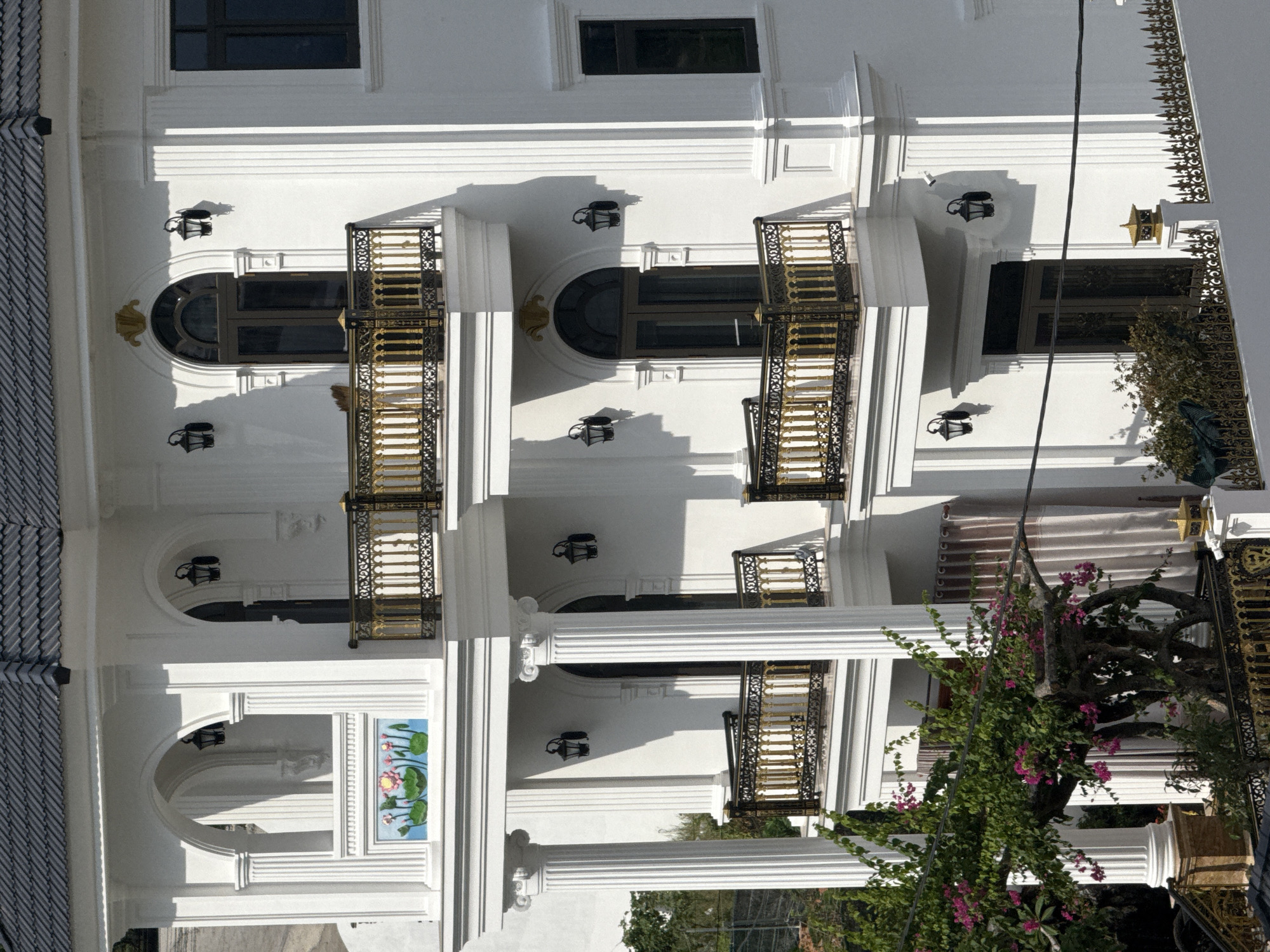 This is a full shot, eye-level, exterior photograph of a white, multi-story house with an ornate architectural style, possibly located in Bac Nha Trang Ward, Vietnam. The house features prominent white columns, arched windows, balconies with gold and black railings, and decorative lighting fixtures. A lush green tree with pink flowers is partially visible in the foreground on the left side of the image. The house has a dark gray tiled roof. The overall impression is one of luxury and traditional design.