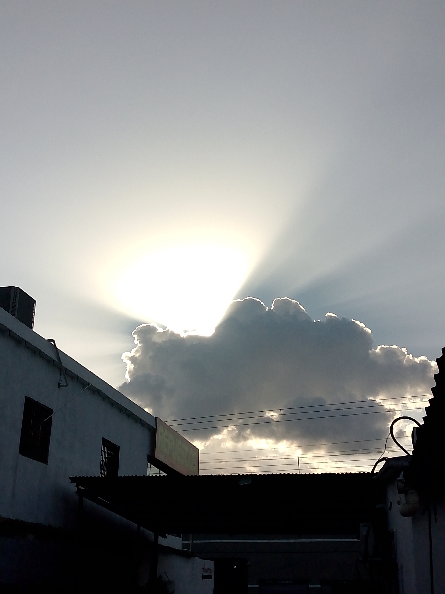 The image captures a bright sky obscured by a large cloud in Tinaquillo, Venezuela. The sun is behind the cloud, creating visible sunbeams radiating outwards. In the foreground, the silhouettes of buildings, rooftops, and power lines are visible, obscuring the view of the city and giving context to the urban location. An air conditioning unit is visible on the roof of one of the buildings.