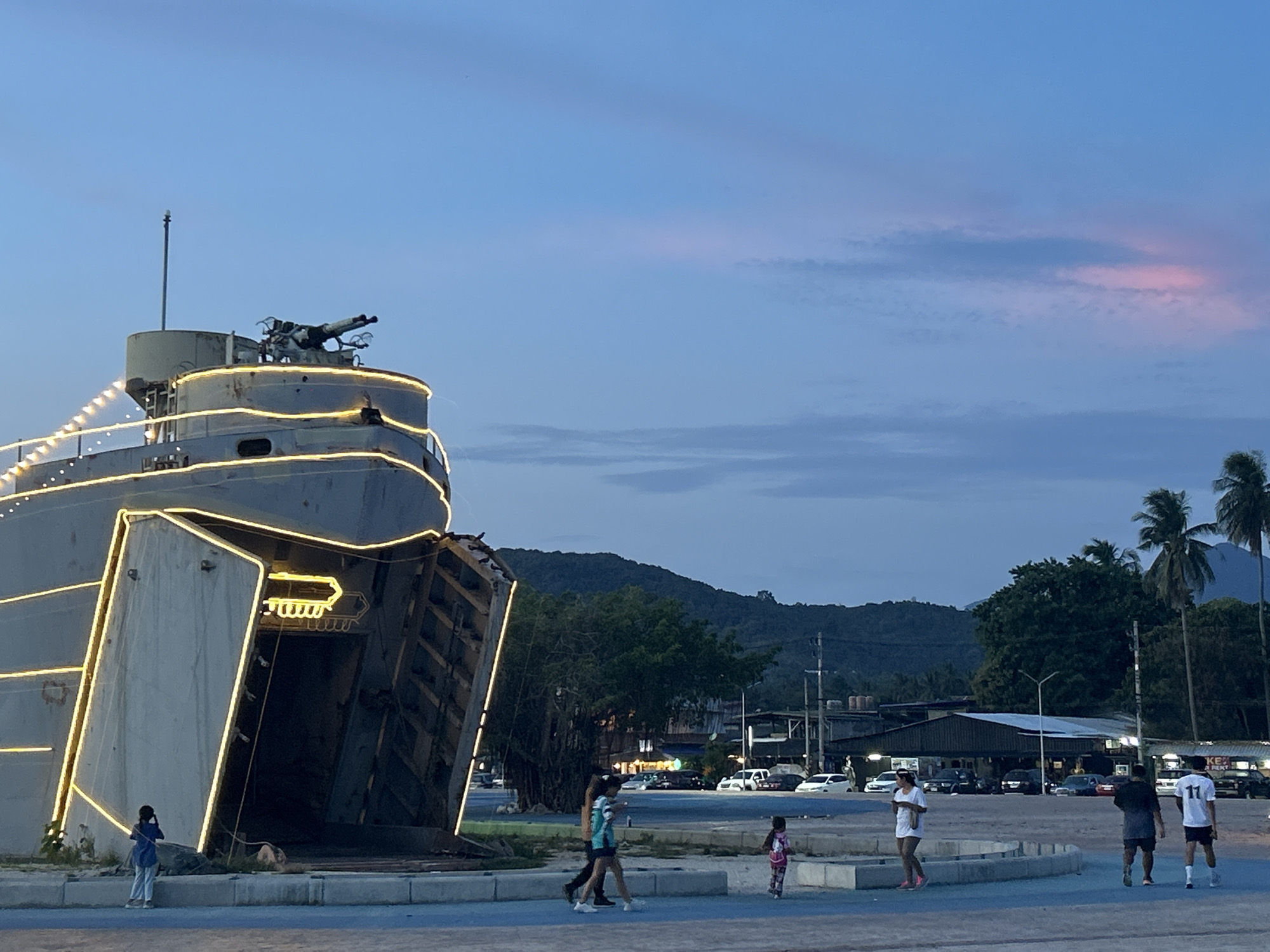 The image shows a partially illuminated structure resembling the front of a ship, decorated with lights, located in Thong Sala, Thailand. A cannon is mounted on the top of the structure. Several people are walking and standing around a blue concrete platform in front of the structure. In the background, there is a mountain range, trees, buildings, parked cars, and a street. The sky is a gradient of blue with some pinkish hues.