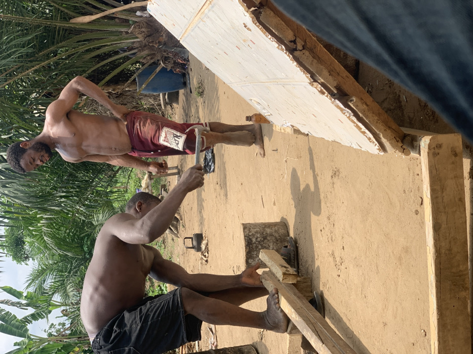 Two shirtless men are working on a wooden structure in Egbeda, Nigeria. One man is using a wrench to tighten a metal bracket. The second man is using a wooden plank to secure the structure. The men are both barefoot and wearing shorts. The structure is being built on a dirt floor and surrounded by vegetation. The location is near the coordinates 7.12 latitude and 5.362 longitude.