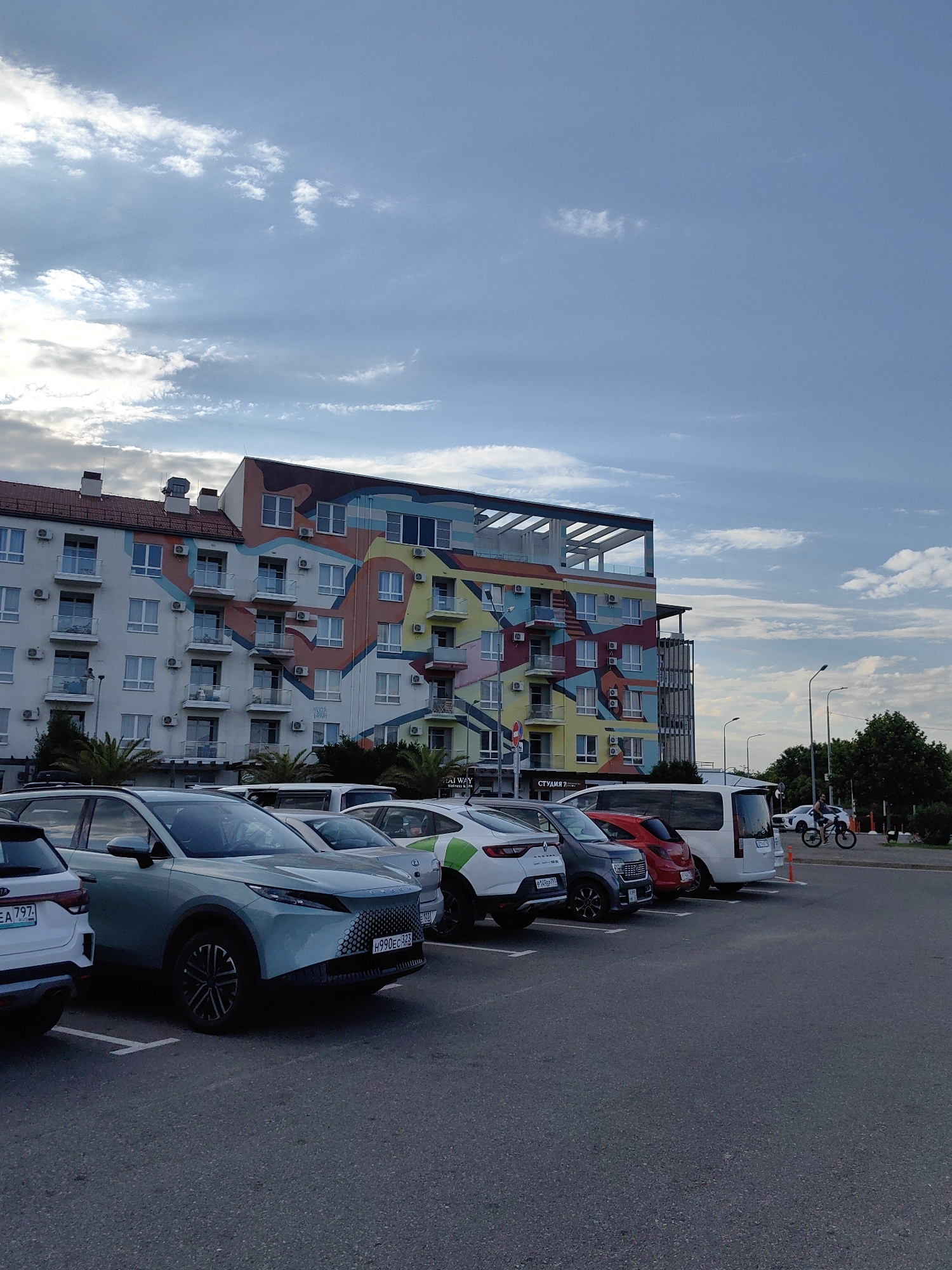 The image shows a parking lot with several cars parked in front of a colorful, modern building in Sirius, Russia. The building features a vibrant mural with geometric shapes and bright colors, including yellow, orange, blue, and pink. The architecture appears contemporary, with multiple balconies and windows. In the foreground, several cars are visible. A light blue SUV is prominently featured in the center, with its front grille and distinctive headlights clearly visible. Other cars, including a white SUV, a dark gray car, and a red car, are parked alongside it. A white van is parked further down the row. In the background, there are streetlights, trees, and a person riding a bicycle. The sky is partly cloudy with a mix of blue sky and white clouds, suggesting it might be late afternoon or early evening. The overall scene depicts a developed urban or suburban area with contemporary architecture and transportation infrastructure.