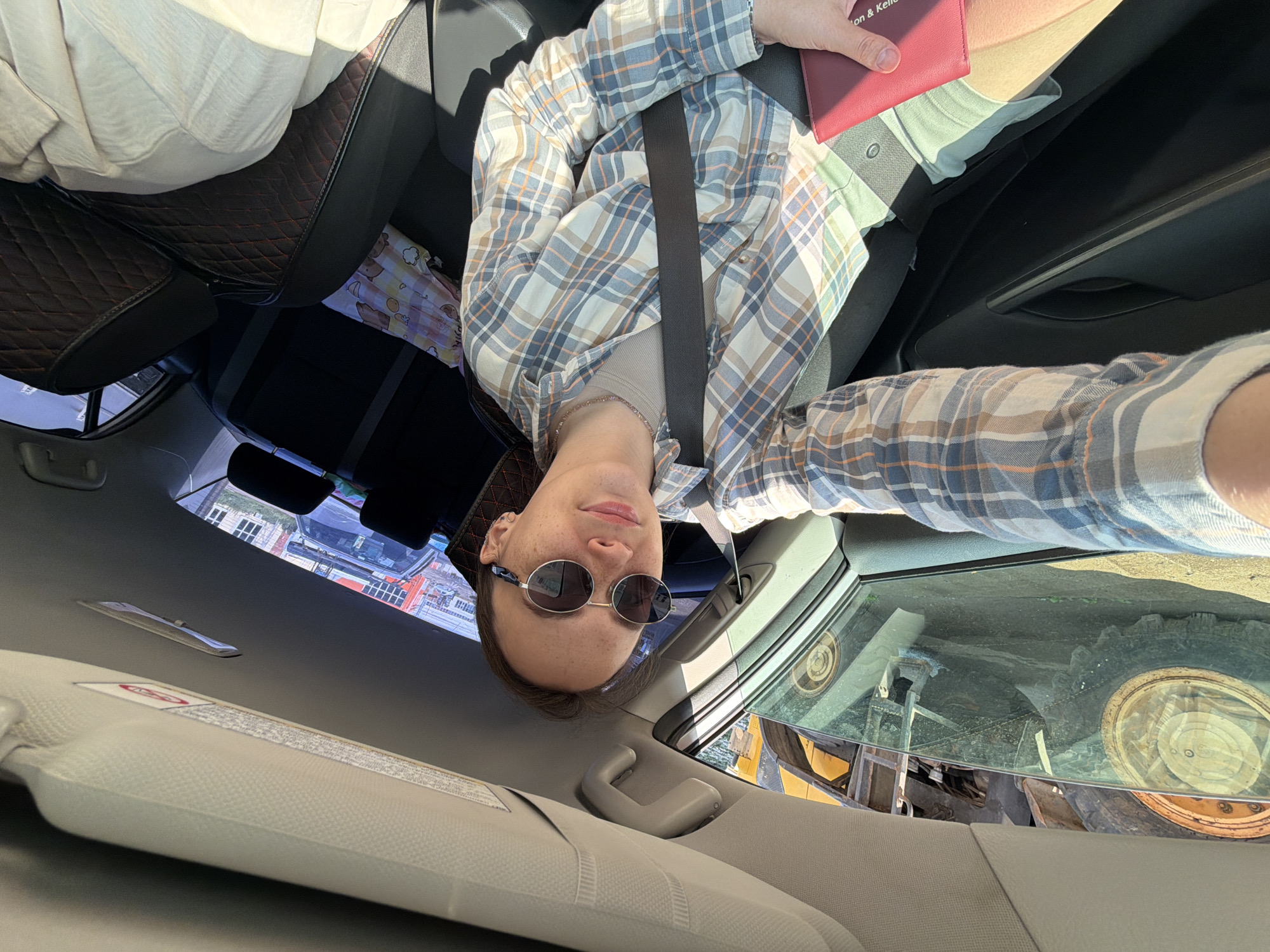 A person is sitting in the driver's seat of a car, holding a red passport, and looking at the camera. They are wearing a plaid shirt and sunglasses. Outside the car window, a large construction vehicle with a yellow arm and a large tire is visible. The car is parked on a dirt surface, possibly a construction site or a rural area. The interior of the car shows car seats and a seatbelt. The background outside the car includes some buildings and what appears to be a road. The presence of a construction vehicle and the terrain suggest the location might be in an industrial area or on the outskirts of the city of Chaikovsky, Russia, where construction and industrial activities are common.