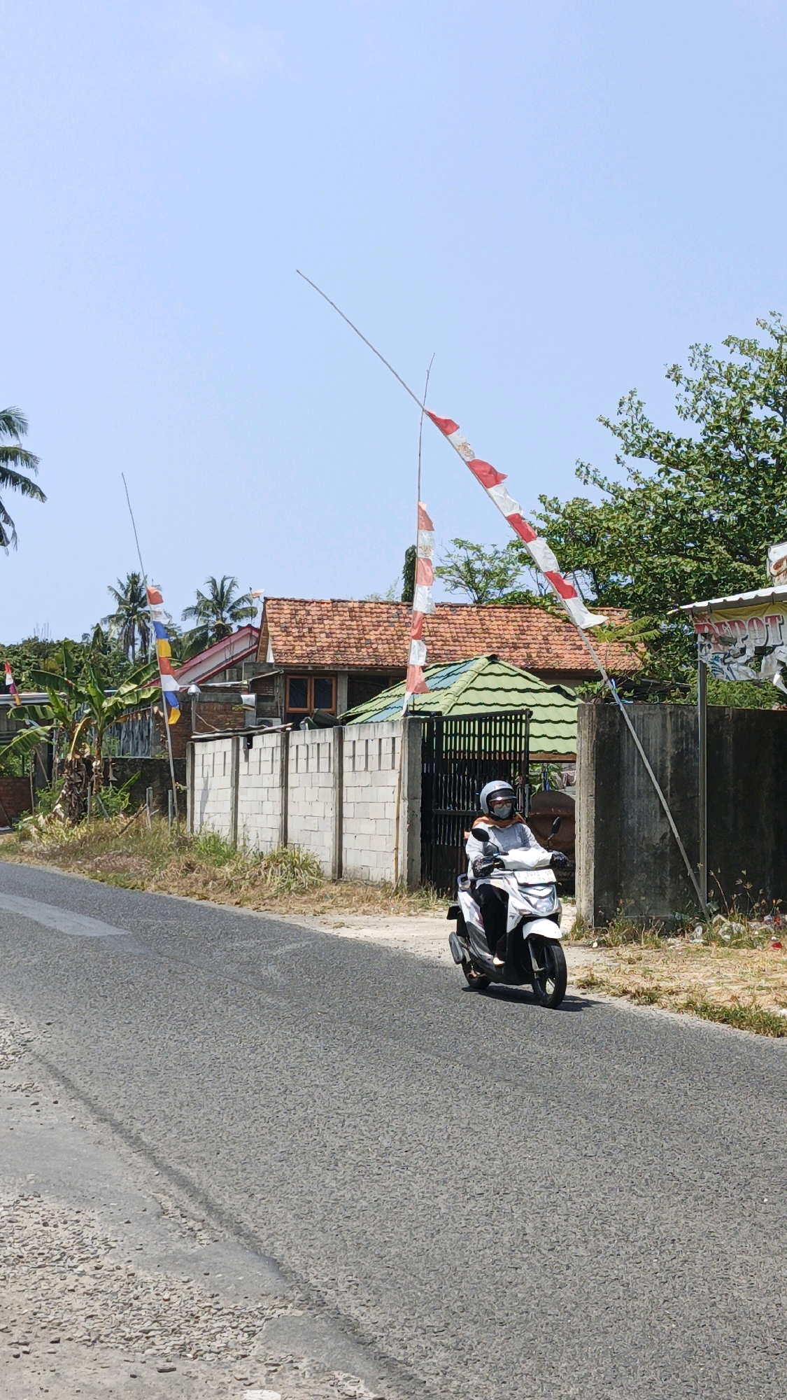 A person rides a white scooter down a paved road in Desa Karangjaladri, Indonesia. The person is wearing a helmet, a light gray long-sleeved shirt, and dark pants. The scooter is moving towards the right side of the frame. On the right side of the road, there is a concrete wall, a black metal gate, a building with a green roof and a building with a red tiled roof. Red and white Indonesian flags are flying from tall poles in front of the buildings. Palm trees and other vegetation are visible in the background. The sky is clear and blue. The road has some rough patches and gravel on the sides.