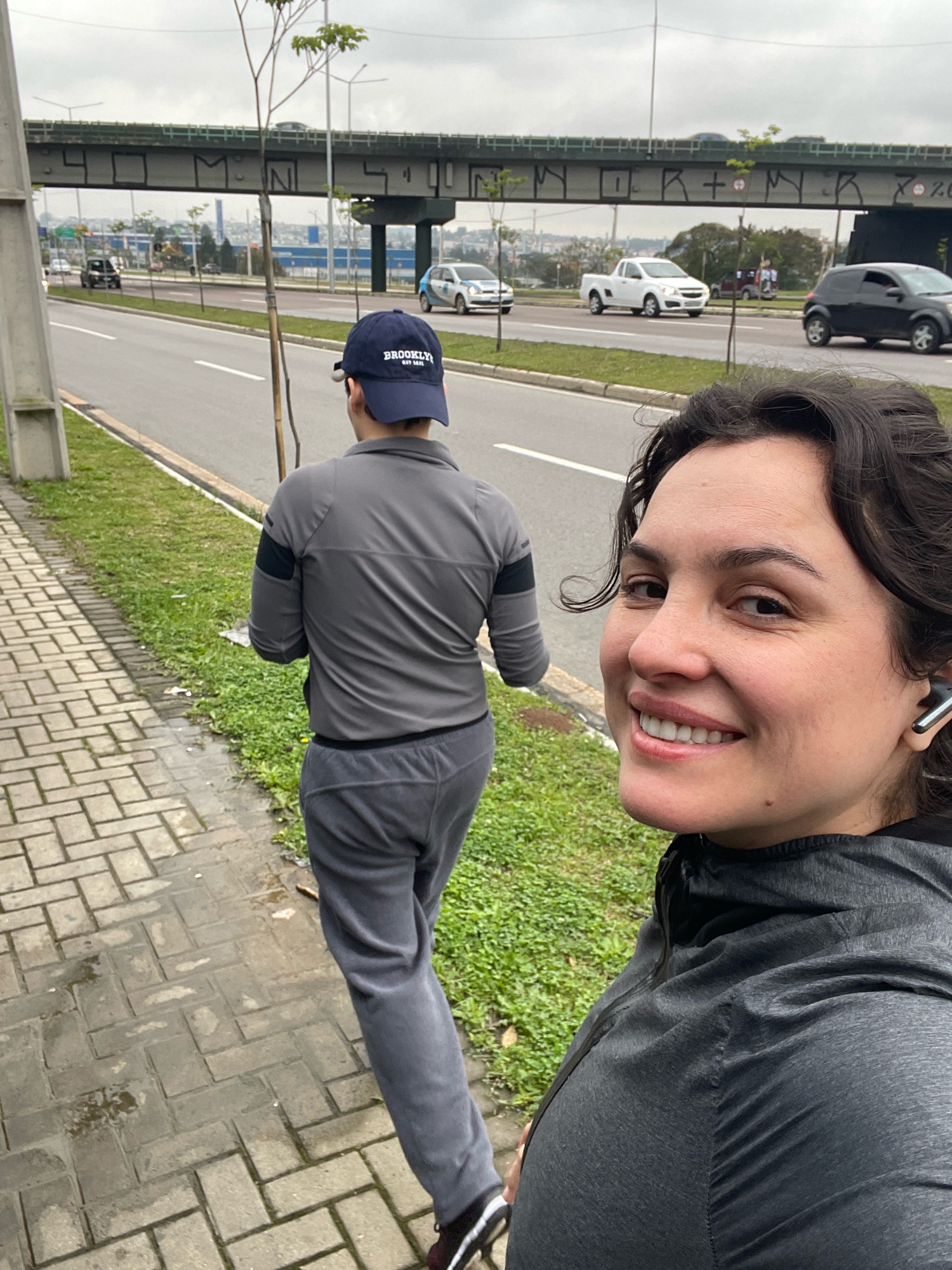 Two individuals are walking on a brick sidewalk adjacent to a multi-lane road. A person in the foreground, facing the camera, is smiling and wearing a dark gray hooded jacket and wireless earbuds. To their left and slightly behind, another person is walking away from the camera, wearing a gray jacket and a blue baseball cap with "BROOKLYN" written on it. They are both on the sidewalk, with a grassy area separating them from the road. Cars and a white pickup truck are visible on the road, and a pedestrian overpass with graffiti is in the background. The environment appears to be urban, with a cloudy sky. The scene is likely in the city of Curitiba, Brazil, given the context of previous images.