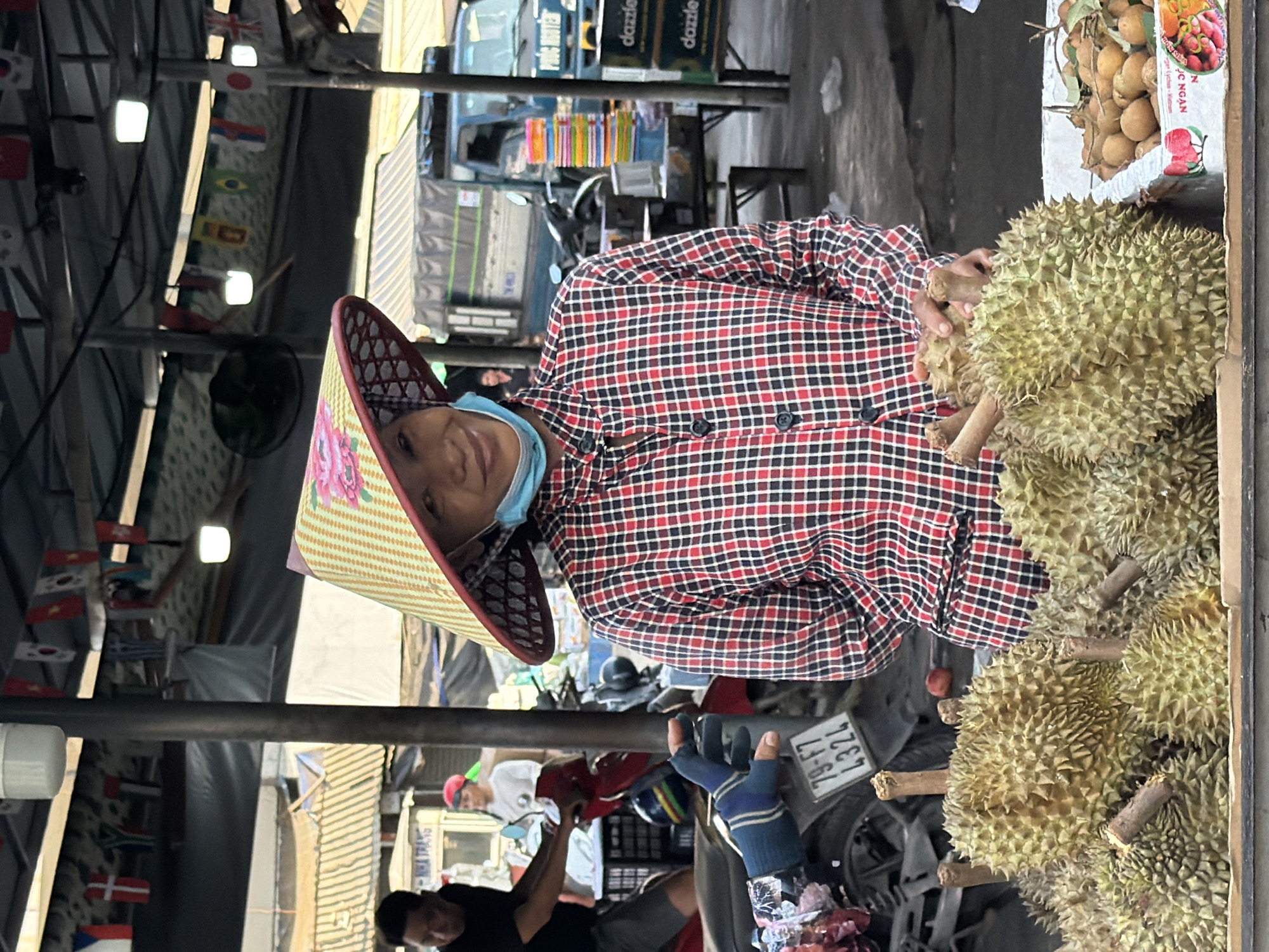 A woman wearing a conical hat and a face mask smiles at the camera while standing in front of a market stall in Nha Trang, Vietnam. She is wearing a red and black checkered shirt. In front of her, there are several durians in a wooden crate. In the background, there are other market stalls, a blue truck, and motorcycles. The overall scene depicts a typical street market in Nha Trang.