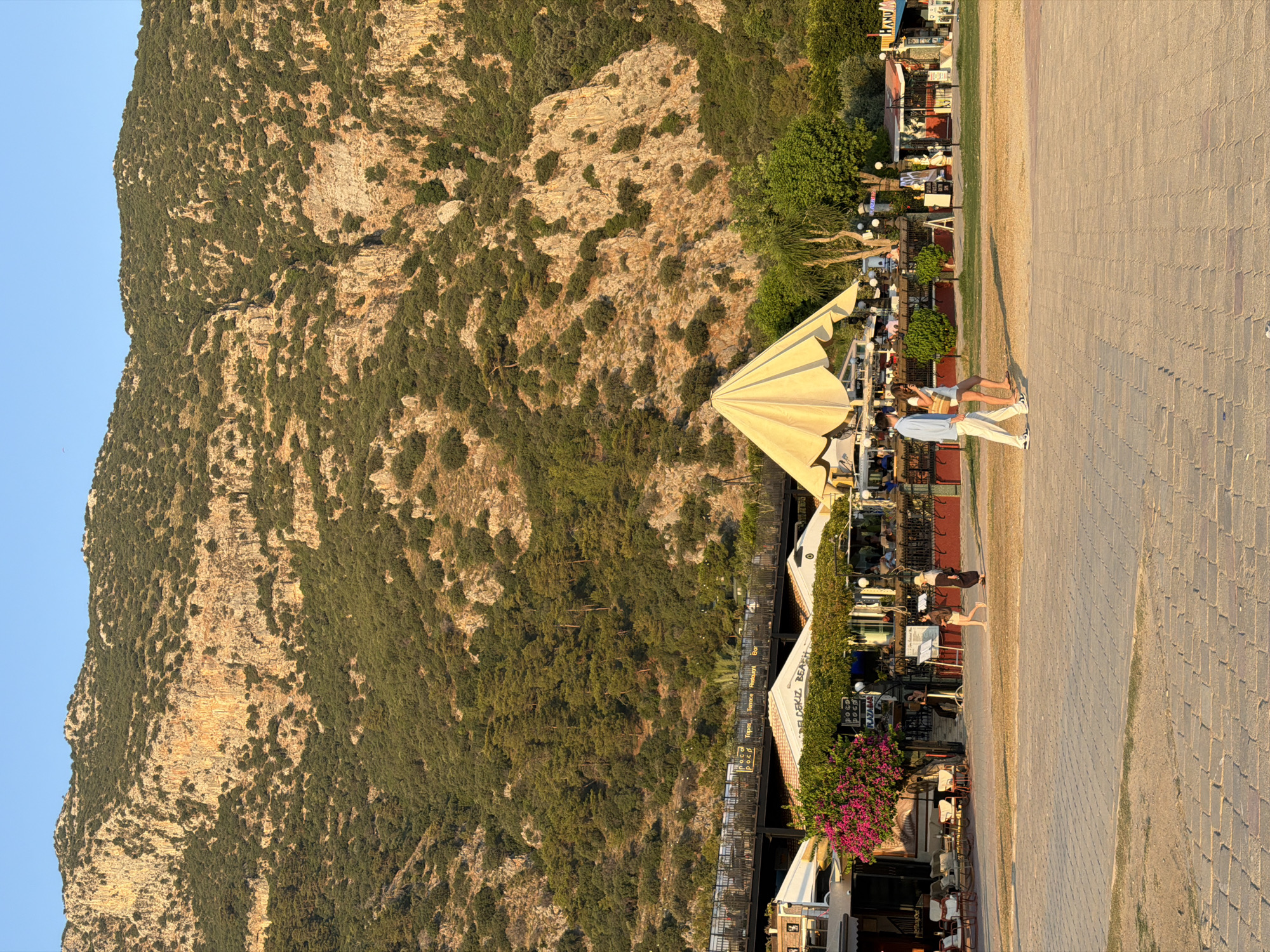 The image depicts a sun-drenched scene in Fethiye, Turkey, likely at Ölüdeniz beach, with a prominent, tree-covered mountain dominating the background. In the midground, a row of businesses, including restaurants and shops, is visible, some featuring outdoor seating and awnings. A large, pale yellow umbrella is a central element among the establishments. Several people are walking on a paved area in front of these businesses. In the foreground, the ground is paved with large, rectangular stones. The overall impression is of a vibrant, seaside leisure area on a clear day.