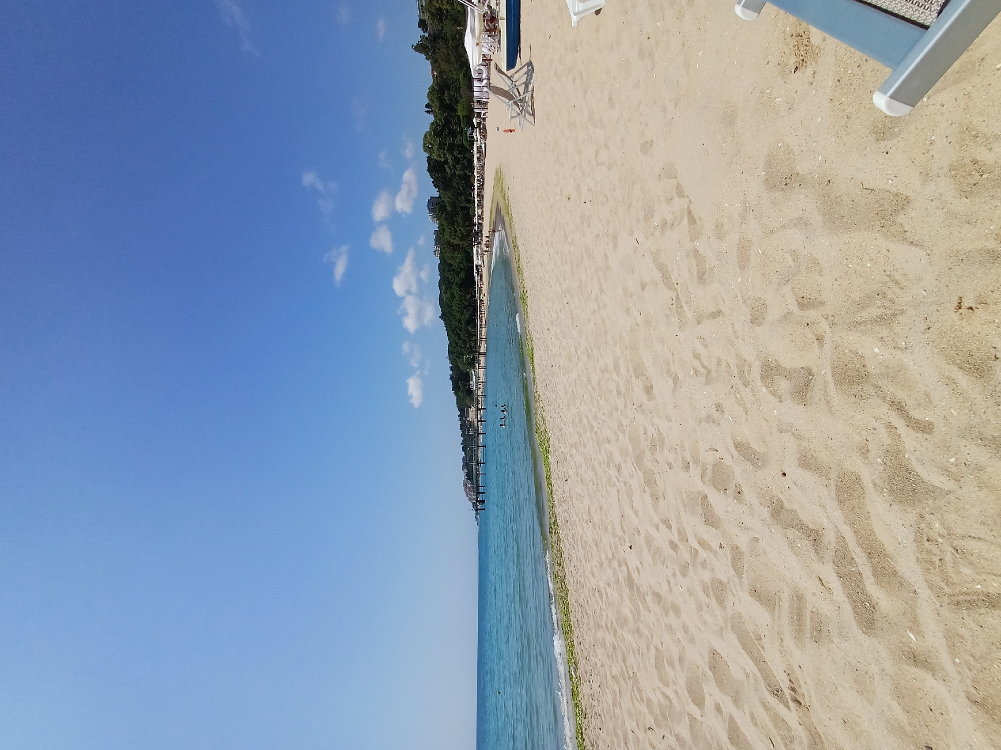 The image depicts a tranquil beach scene in Varna, Bulgaria, likely in the northern part of the city given the presence of a pier and resort buildings along the coastline. The foreground is dominated by a wide expanse of sandy beach, marked with footprints and scattered shells. In the mid-ground, the calm, turquoise waters of the Black Sea gently lap the shore, with a line of seaweed visible along the waterline. A few people are visible wading and swimming in the shallow water.
Further in the distance, a long pier extends into the sea, serving as a walkway and likely a recreational spot. Along the coastline, a strip of green trees forms a backdrop to the beach, interspersed with resort buildings and white umbrellas, suggesting a developed tourist area. The sky above is a clear, vibrant blue, with a few wispy white clouds adding texture. Two empty white lounge chairs are positioned on the sand, hinting at a relaxing day at the beach.