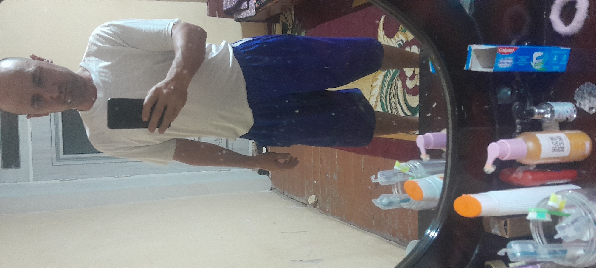 A bald man in a white t-shirt and blue shorts takes a selfie in a mirror. He is holding a black smartphone in his right hand. The mirror reflects a room with a wooden floor and a beige wall. There is a red and yellow patterned rug on the floor. In the foreground, reflected in the mirror, is a dark wooden vanity with several toiletries, including toothbrushes in a jar, a white bottle with an orange cap, a pink pump bottle, and a blue toothpaste box. A bed with a patterned cover is visible in the background to the right. The image appears to be taken indoors, likely in a bathroom or bedroom.