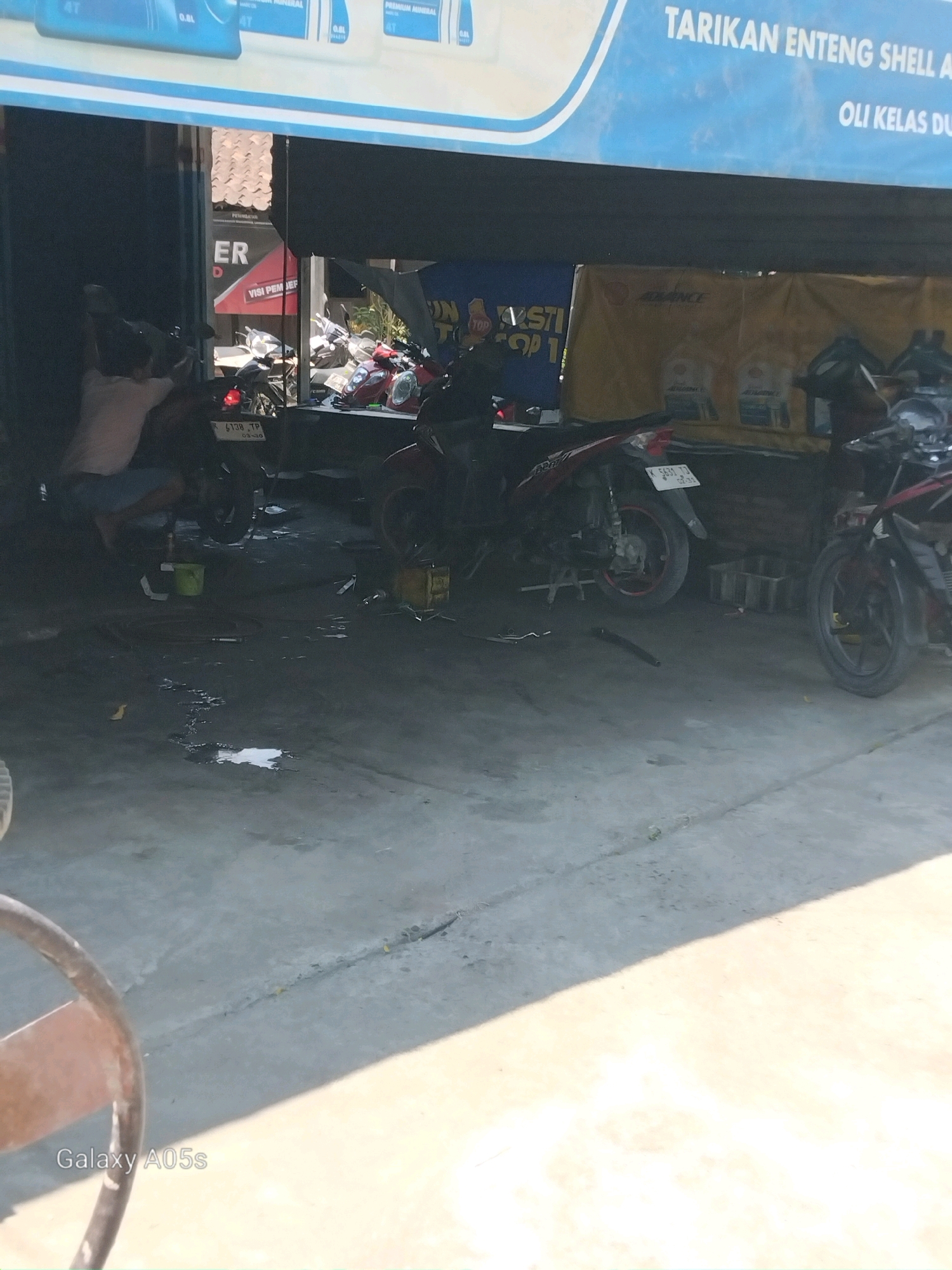 The image depicts an outdoor motorcycle repair shop in Karangayung, Indonesia. The foreground shows a concrete floor with some oil stains and a section of a metal stand. In the middle ground, several motorcycles are parked. One motorcycle is red and black, facing left, with its front wheel obscured. Another red motorcycle is parked in the center, facing left, on a stand. A person, partially visible from the back and side, is crouching next to this motorcycle, appearing to be working on it. This individual is wearing a light-colored t-shirt and dark shorts. To the right, another red and black motorcycle is parked facing right. The background features a large blue banner with white text and a yellow tarp. The blue banner has text that appears to read "TARIKAN ENTENG SHELL A" and "OLI KELAS DUA". The yellow tarp has some text visible, including "ADVANCE". A dark doorway or opening is visible to the left, with a sign that has red and white lettering, including the word "ER". The overall environment suggests a working repair shop with tools and equipment likely present but not clearly visible.