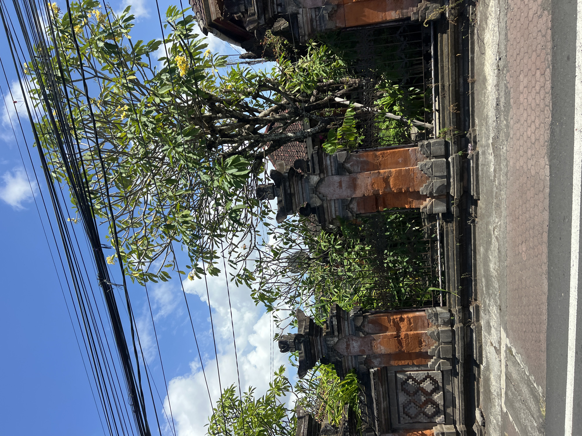 This is a street-level view of a traditional Balinese gate and wall in Denpasar, Indonesia. The gate and wall are made of carved stone and brick, with intricate detailing. A lush green tree with yellow flowers is growing behind the gate, and its branches extend over the wall and into the foreground. Overhead, a tangle of black electrical wires crisscrosses the bright blue sky, which is dotted with fluffy white clouds. The street in front of the gate is paved with reddish-brown interlocking bricks, and a white line marks the edge of the road. The scene appears to be in a residential or temple area, given the architectural style of the gate. There are no people visible in the image.