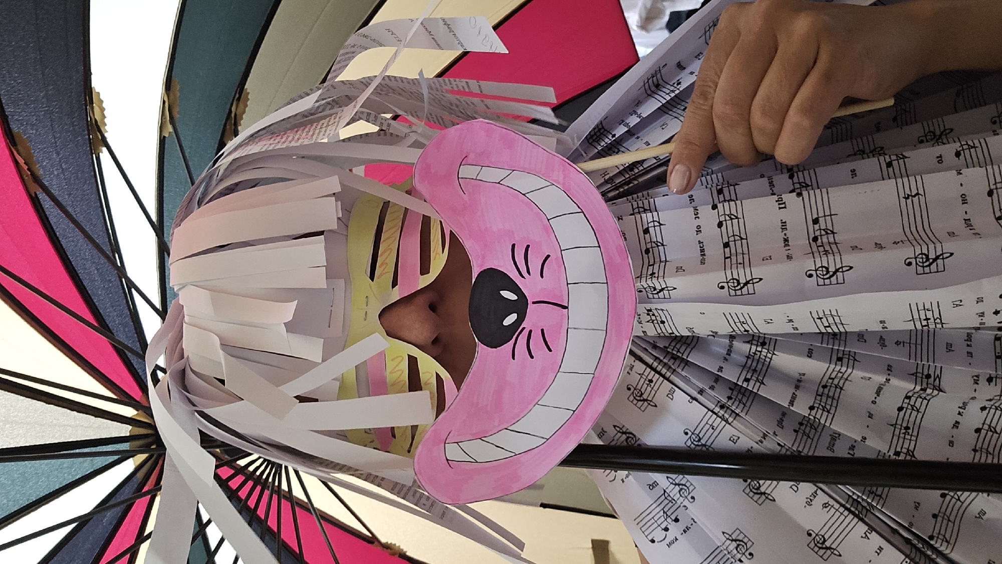 The photograph shows a person wearing a carnival costume. The person's face is partially visible, with their nose visible behind a pink mask depicting a smiling cat. The costume includes strips of white and yellow paper serving as a wig. The mask is attached to a thin, wooden stick held by a hand, which also seems to be manipulating other parts of the costume. The costume includes paper sheets with musical notes, folded and arranged to create a fan effect around the person's body. In the background is an open umbrella with panels of pink, blue, and white.