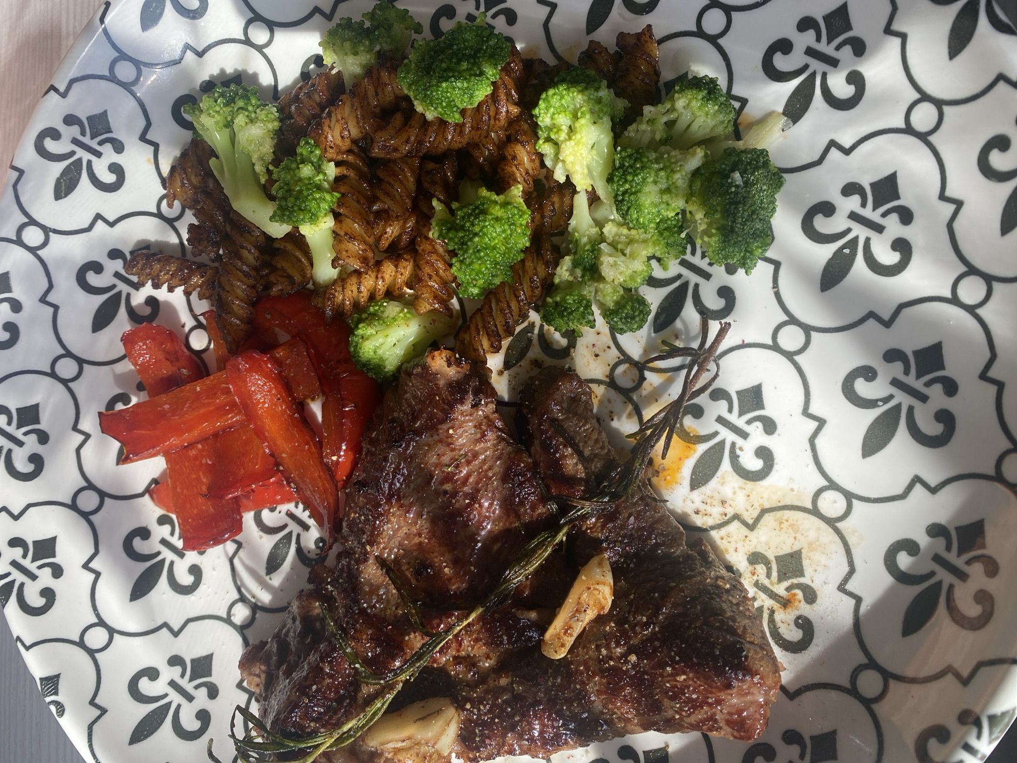 This media file displays a close-up, overhead view of a plated meal. The primary subject is a serving of food arranged on a white plate with a black and gray decorative pattern.
The meal consists of several components:
* A substantial piece of grilled or pan-seared steak, seasoned with what appears to be salt and pepper, occupies the lower right portion of the plate. It is garnished with fresh rosemary sprigs and a few cloves of roasted garlic.
* Next to the steak, in the lower left, are several strips of roasted red bell pepper, showing some charring on the edges.
* Filling the top half of the plate is a portion of dark-colored pasta, likely a rotini or fusilli shape, mixed with florets of steamed or lightly cooked broccoli. The pasta appears to be coated in a sauce or seasoning.
The plate itself is white with an intricate, repeating pattern of black and gray stylized floral or geometric motifs. Strong, distinct shadows are cast across the plate from items outside the frame, indicating bright, direct lighting, possibly natural sunlight coming from a window. There is no visible text in the image.
The image captures a prepared meal, suggesting an activity of eating, likely lunch or dinner, in what appears to be an indoor setting due to the distinct shadow patterns. The location context provided is Chaikovsky, Russia.