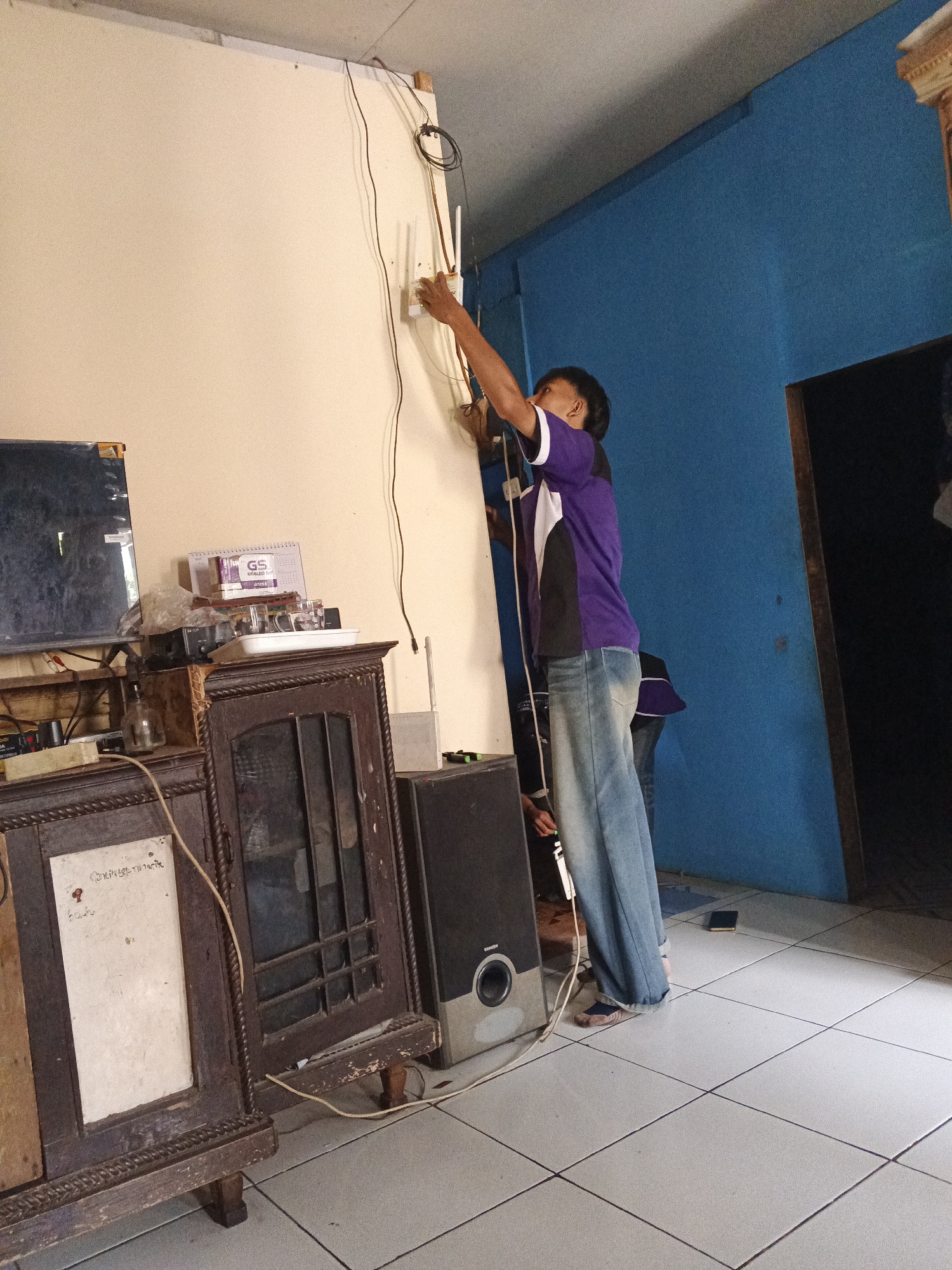A man in a purple and white shirt and blue jeans is installing a white Wi-Fi router on a cream-colored wall. He is standing on a tile floor in a room with blue walls. To the left of the man, there is a wooden cabinet with a glass-fronted door, and on top of the cabinet are various items, including a television, a calendar, and some small containers. A speaker is placed on the floor to the right of the cabinet. Another person, wearing a purple shirt and jeans, is partially visible behind the first man. The scene appears to be an indoor residential setting in Warungkiara, Indonesia.