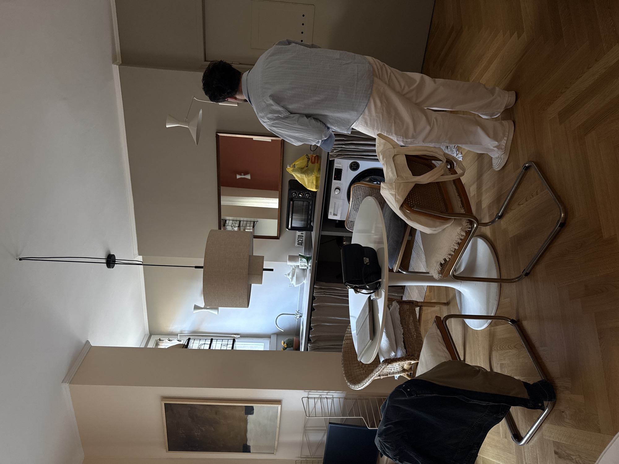 A medium-shot photograph taken from a slightly elevated perspective captures the interior of what appears to be an apartment or a modern home. The individual standing, likely a resident or guest, has dark hair. The person is dressed in a light blue striped shirt and white pants, suggesting a casual or relaxed setting. A washing machine and kitchen appliances are visible. The dining area features a round white table surrounded by chairs of mixed styles: some with woven elements and others with metal frames. A lamp hangs above, providing illumination. A painting on the wall is partially visible, adding to the interior decor. The photograph’s location is in Milan, Italy.
