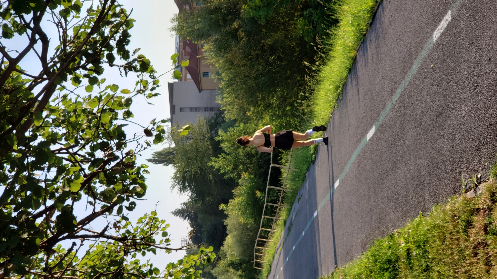 A woman runs on a paved path in Prešov, Slovakia, bordered by green grass and a yellow metal fence. She is wearing a black sports bra and black shorts with white socks and black athletic shoes. Her hair is pulled back in a bun. Trees with green leaves hang over the path, casting shadows. In the background, there are buildings and more trees. The sun is shining, indicating it is likely daytime.