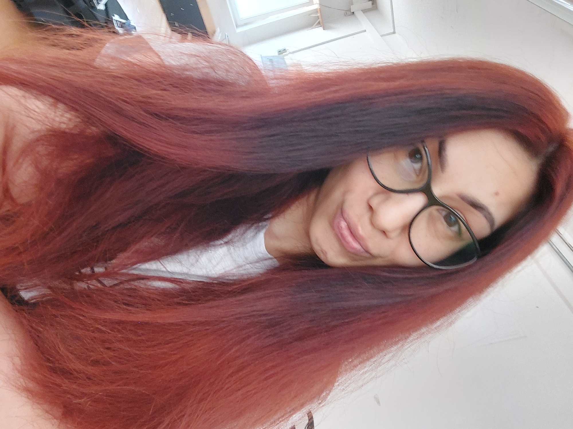 The image is a close-up selfie of a woman with long, reddish-brown hair and glasses. She is wearing a grey t-shirt. Her lips are slightly pursed. The background appears to be an indoor setting with a white wall and a window on the left side. There is no indication of the city of Landshut, Germany, or any specific location within it. The image is focused on the woman's face and upper body.