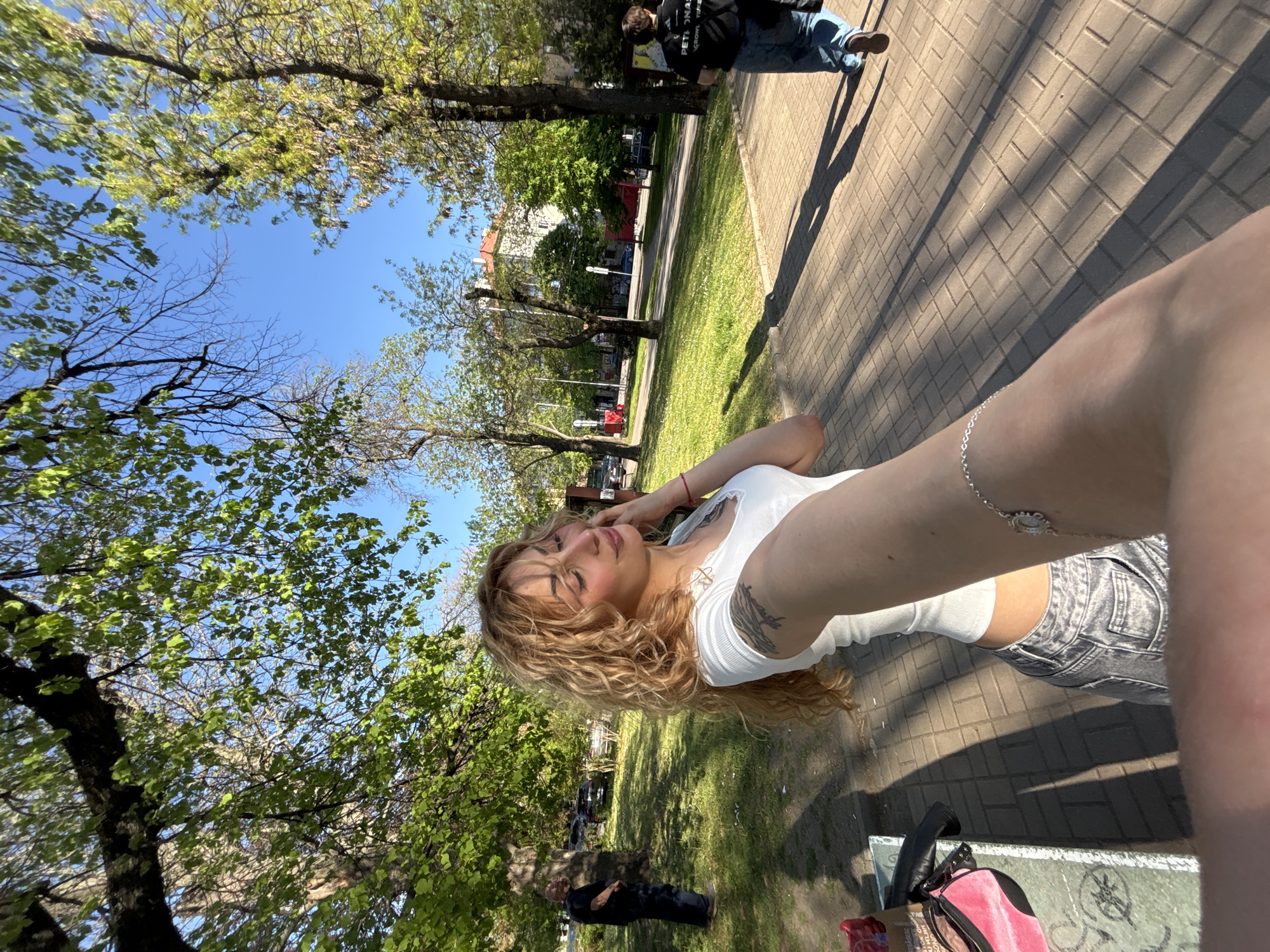 A woman with blonde curly hair, wearing a white top and gray shorts, takes a selfie outdoors in a park. She is fair-skinned with tattoos on her left arm. A man in a black jacket and blue jeans stands on a paved pathway nearby. Trees with green leaves surround the area, and a patch of grass is visible between the pathway and the trees. There is a clear blue sky above. In the background, a building with a red roof is seen. The park appears to be located in an urban area of Sofia, Bulgaria. Additional items such as a pink bag and a possibly a bench are visible near the selfie taker.