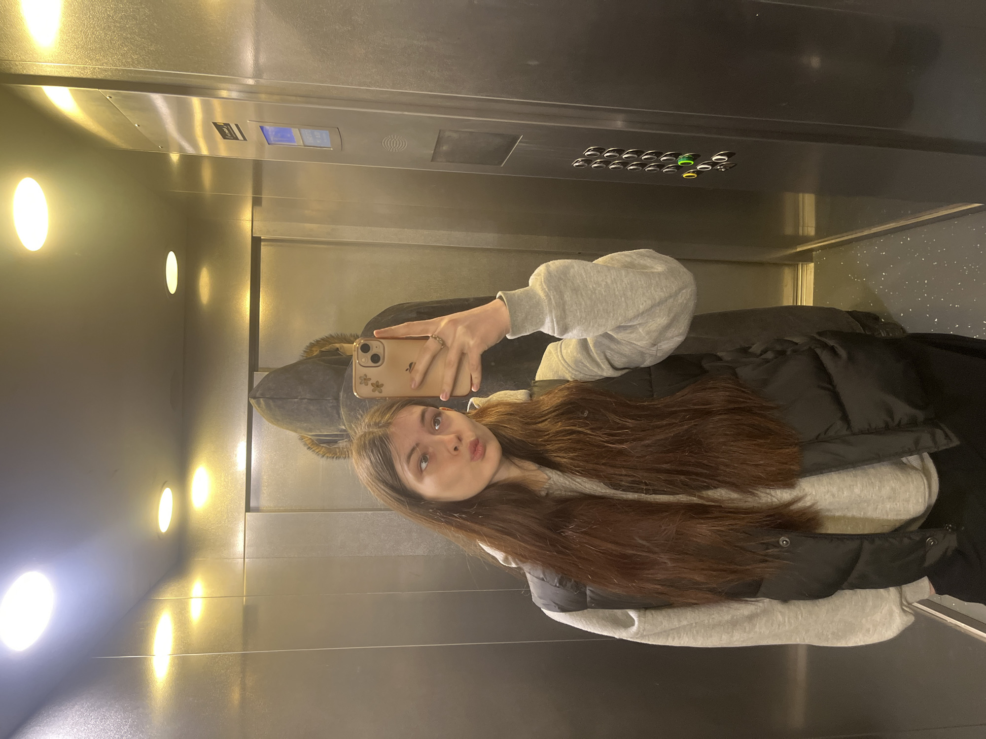 A young woman is taking a selfie in an elevator. She is wearing a gray hooded sweatshirt and a black puffer vest. Her brown hair is long and straight. She is holding a beige smartphone with a flower sticker on the back. The elevator has a mirrored wall with round lights and a control panel with buttons. The floor appears to have a speckled pattern. The location cannot be determined to be Saint Petersburg, Russia from this image.