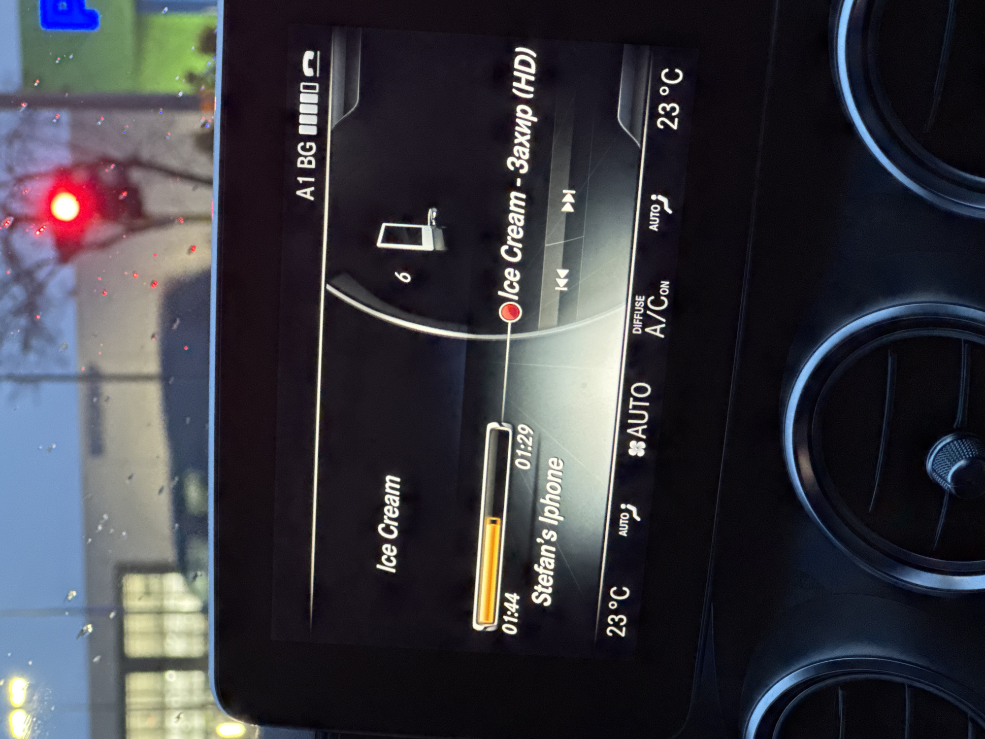 The image shows the entertainment system of a Mercedes-Benz car. The screen displays music information. The song "Ice Cream - Захир (HD)" is playing from "Stefan's Iphone." The song is 01:44 into a 01:29 song. The car's climate control is set to "AUTO" and the temperature is 23°C. The phone signal is full, and the phone is connected to the car. "A1 BG" is visible, indicating that the network provider is A1 Bulgaria. The car is likely in Sofia, Bulgaria. A blurred view outside the car window reveals a traffic light and other buildings. It is raining.