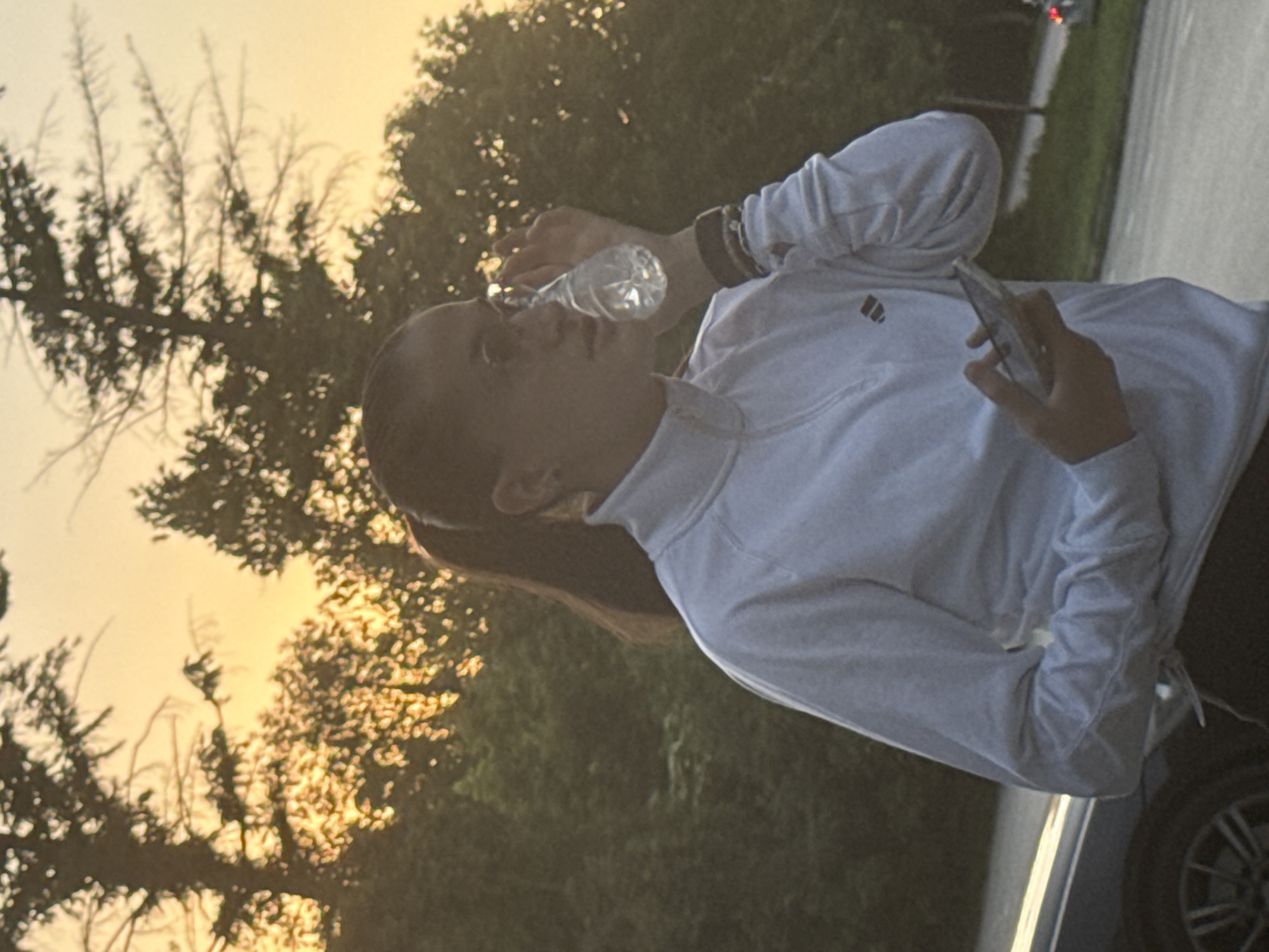 A person, likely female, is standing outdoors, holding a smartphone in their right hand and a bottle of water up to their mouth with their left hand. The individual is wearing a white long-sleeved shirt or sweatshirt and dark leggings. The background consists of trees and the sky, possibly at sunset, with some light filtering through the branches. A car is partially visible in the lower right corner of the image. The image does not show any visual cues that indicate it was taken in the city of Pleven, Bulgaria.