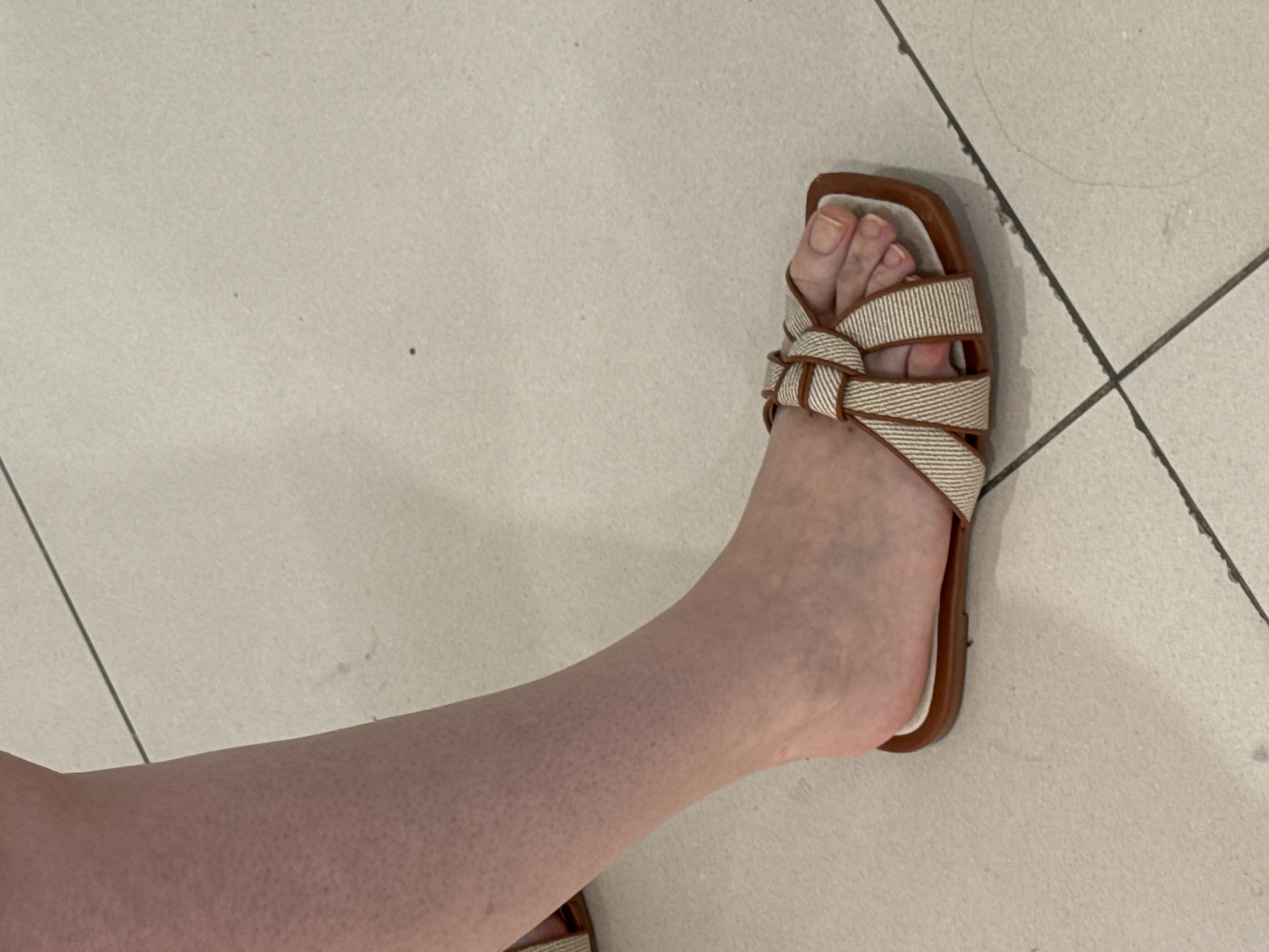 A fair-skinned person's lower left leg and foot are shown from a slightly elevated perspective, resting on a tiled floor. The skin on the lower leg and foot exhibits a texture consistent with small goosebumps or piloerection. A faint reddish mark is visible on the inner calf. The foot wears an open-toed, flat sandal with a square-shaped sole. The sandal's upper features two crisscrossing straps made of a woven, beige-and-brown striped fabric, knotted at the center. The sandal's footbed is white, and its sole is a solid brown. The visible toenails are natural, unpainted, and appear slightly long. The floor consists of light-colored, possibly off-white or light grey, rectangular tiles separated by thin, dark grout lines. Small dark specks and a thin strand of dark hair are present on the tiled surface. The lighting is uniform, suggesting an indoor environment. The image does not contain information to identify a specific location in Plovdiv, Bulgaria.