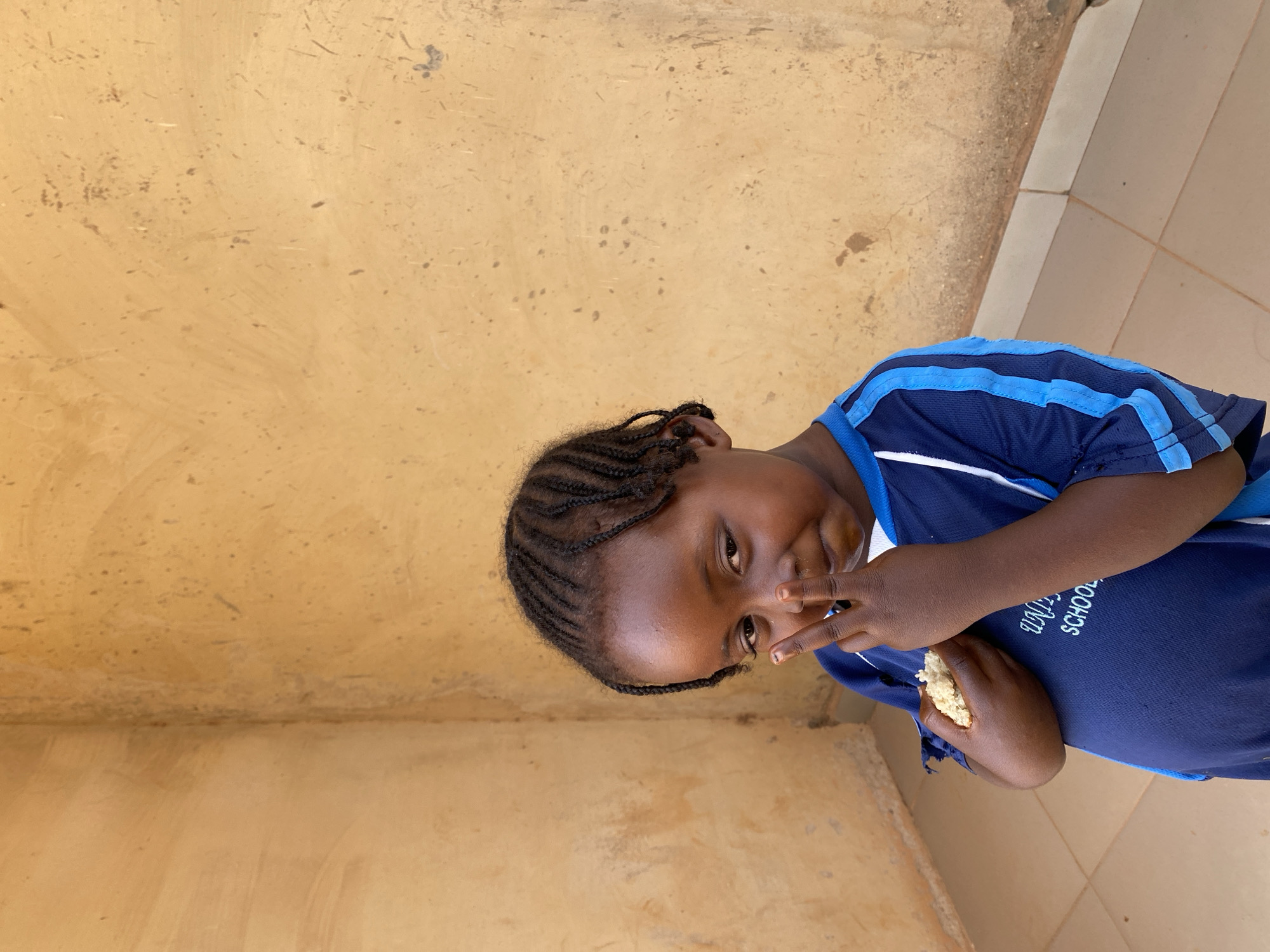 A young, dark-skinned child with braided hair is holding food and posing for the camera near a wall. The child wears a blue shirt with blue stripes on the shoulders and a logo that reads "WOODEN SCHOOL" in script. The wall is a muted yellow with visible imperfections, suggesting an outdoor setting. The scene is located in Kuru Dungus, Nigeria.