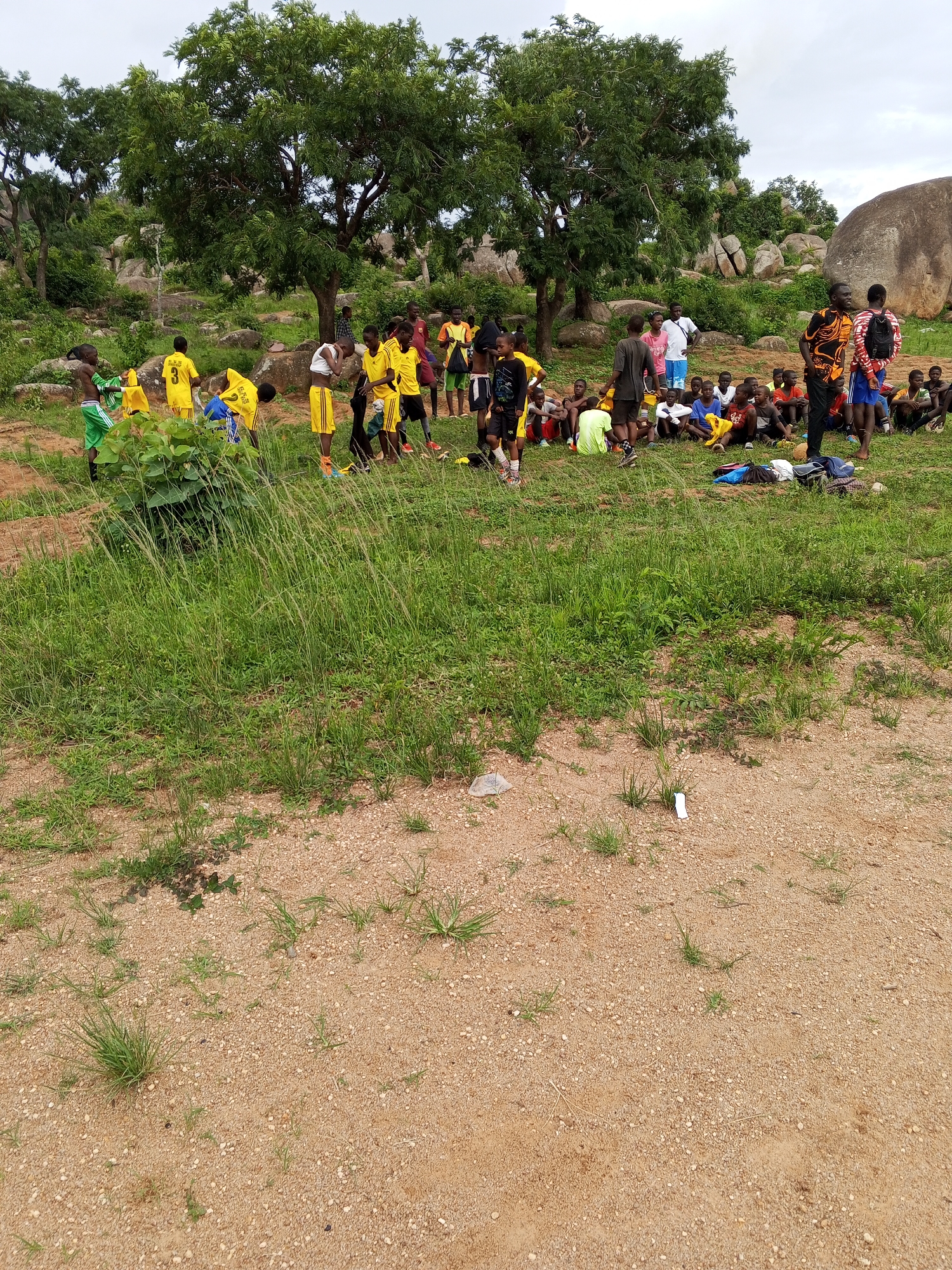 The image depicts an outdoor scene in Gwong, Nigeria, with a group of young people, primarily boys, gathered in a grassy area with a sandy path in the foreground. The environment is characterized by large, rounded rocks and lush green trees with dense foliage, suggesting a somewhat rugged or natural landscape. The sky is overcast.
In the foreground, the sandy path is sparsely covered with dry grass and small pebbles. Moving towards the middle ground, a patch of tall, green grass dominates the scene. A group of about thirty to forty individuals, mostly young boys, are visible. Many of them are wearing athletic attire, predominantly yellow and green shirts and shorts, with some in blue. Some individuals are standing, while others are sitting on the ground, appearing to be resting or preparing for an activity. A few adults are also present, one wearing a colorful patterned shirt and another in a white shirt.
Several objects are scattered on the ground, including bags, some appearing to be sports bags, and possibly towels or clothing items. The overall impression is a gathering for a sports event or practice session in a somewhat informal setting. The natural elements, including the rocks and trees, are prominent and form a backdrop to the human activity.