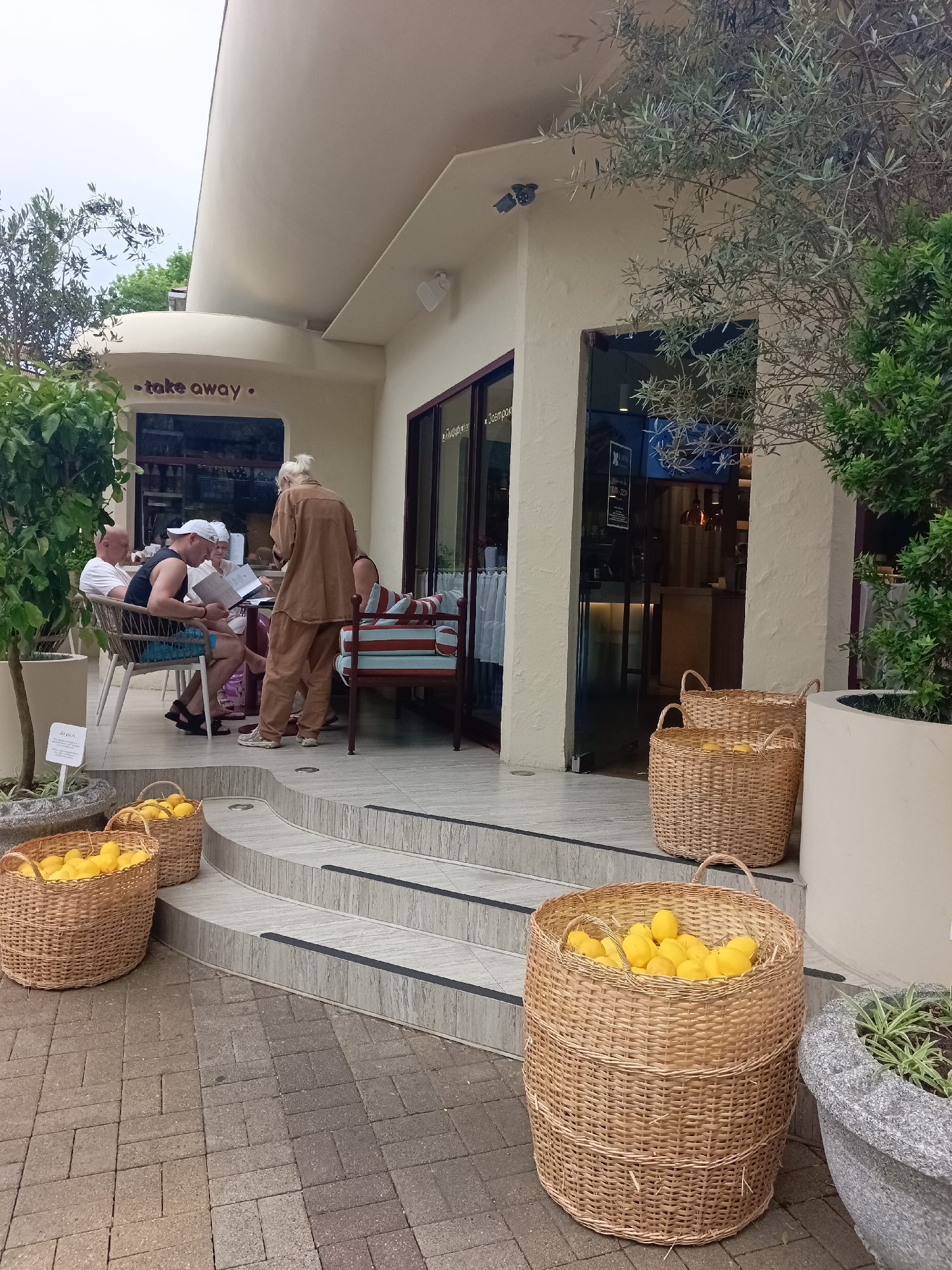 In Sochi, Russia, an outdoor seating area of a restaurant is depicted. Three men and a woman are seated at a table, appearing to be engaged in conversation or looking at menus. Another woman stands, facing the group. Baskets filled with lemons are placed on the ground and steps leading up to the restaurant's entrance. The restaurant's facade is visible, with "take away" signage above an entrance. An olive tree and other greenery add to the scene's ambiance. The weather appears overcast.