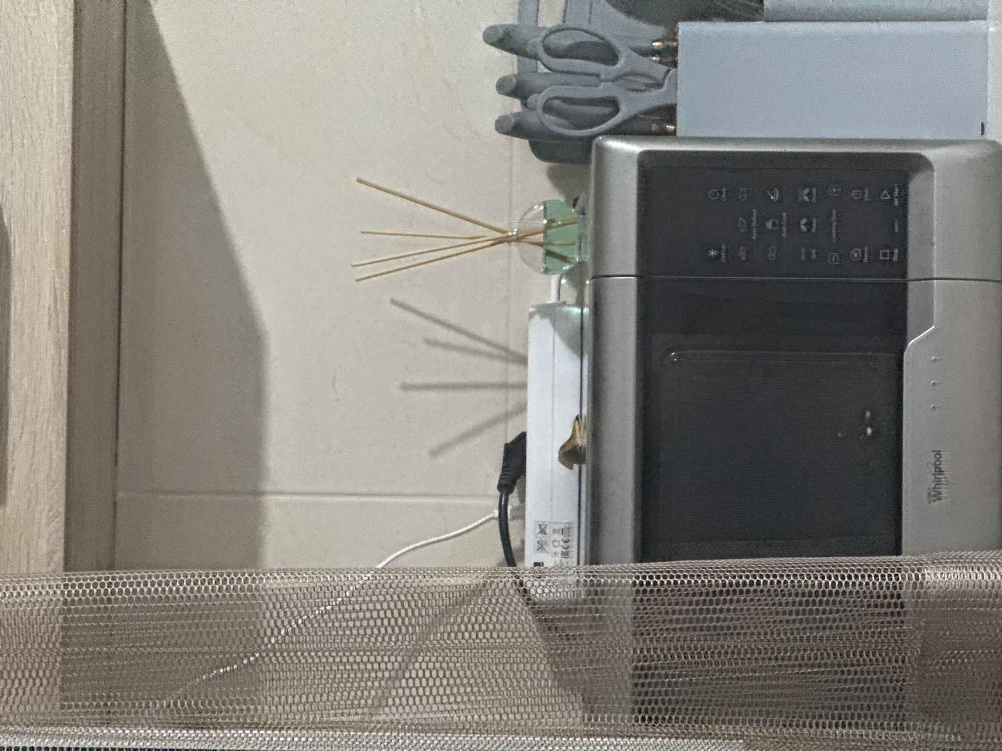 The image captures a close-up view of a kitchen counter in Chișinău, Moldova. On the counter, a silver Whirlpool microwave is prominently displayed. To the right of the microwave, a light blue container holds several pairs of gray scissors, arranged vertically. Behind the microwave and to its left, a white electronic device with a black power cord is partially visible, with a small, light brown object resting on top of it. A sheer, textured curtain in a neutral tone hangs on the left side of the frame, obscuring some of the background. A small, round glass vase containing a light green liquid and several wooden reeds is positioned on the counter between the white electronic device and the scissors. The background consists of a tiled wall and a wooden shelf.