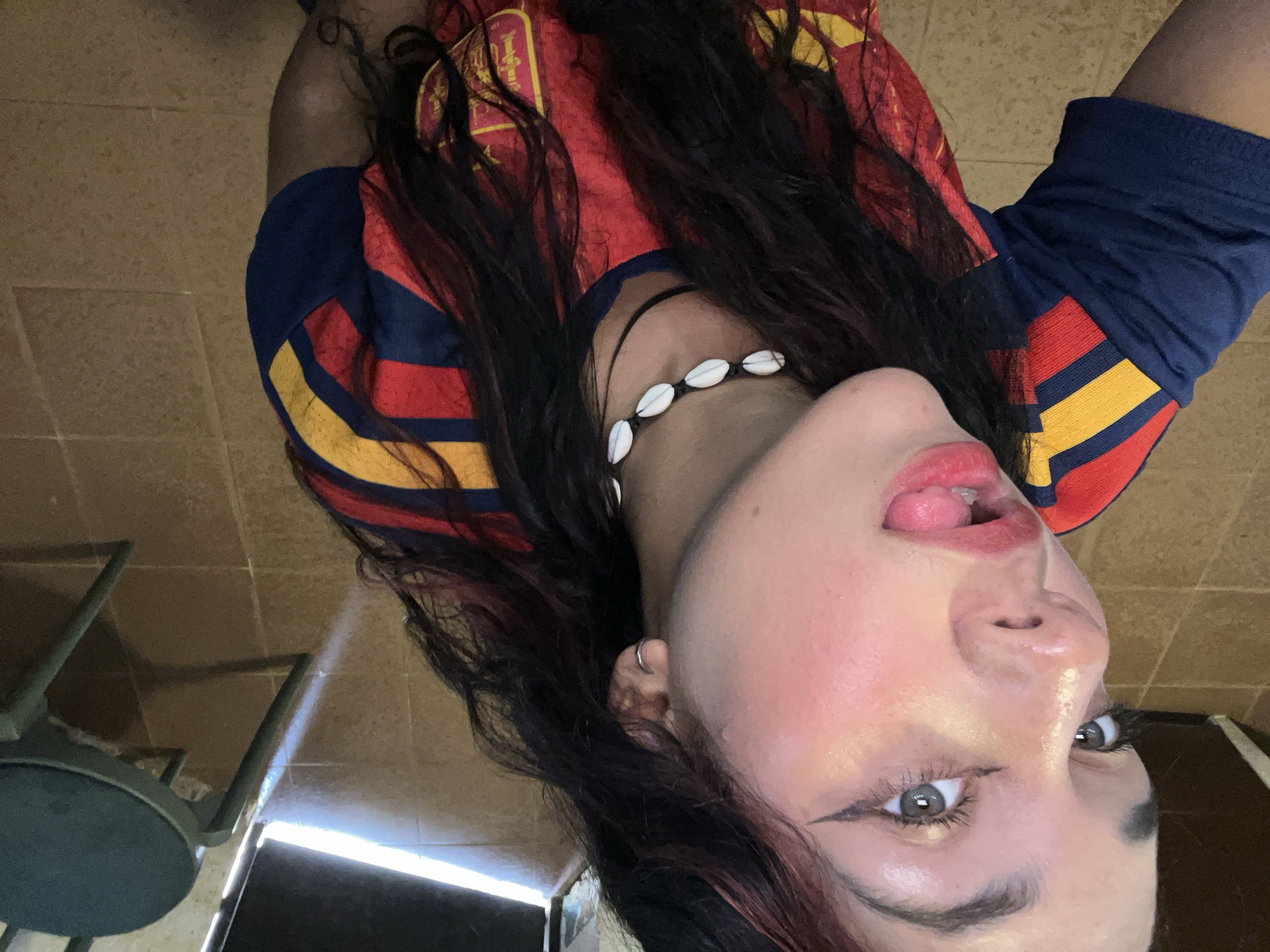 The image shows a close-up, high-angle, selfie-style shot of a young woman with long, dark, wavy hair, some streaks of red visible. She is wearing a blue, red, and yellow striped shirt, possibly a sports jersey. She has on makeup, including winged eyeliner and lipstick. Her tongue is sticking out. She is also wearing a choker necklace made of white shell-like objects. She has a silver hoop earring in her left ear. The background appears to be an indoor space with tiled flooring in shades of brown and beige. A portion of a dark chair with a green cushion is visible to the right. Sunlight streams in from an open doorway or window on the left side of the frame, creating a bright area on the floor and a shadow. The scene is likely within a residential or casual setting. There is no identifiable information within the image to determine its specific location in Porlamar, Venezuela.
