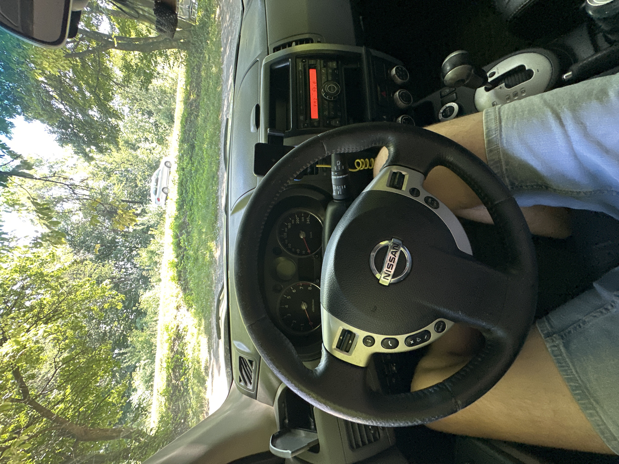 The image is taken from inside a Nissan vehicle, showing the dashboard, steering wheel, and the driver's legs. The driver is wearing denim shorts. The car is stopped on a road bordered by dense green trees and foliage. Sunlight filters through the leaves, creating dappled light. In the distance, a silver car is visible on the road. The radio in the car is on, displaying "FM2 90.8". The location is Shatava, Ukraine.