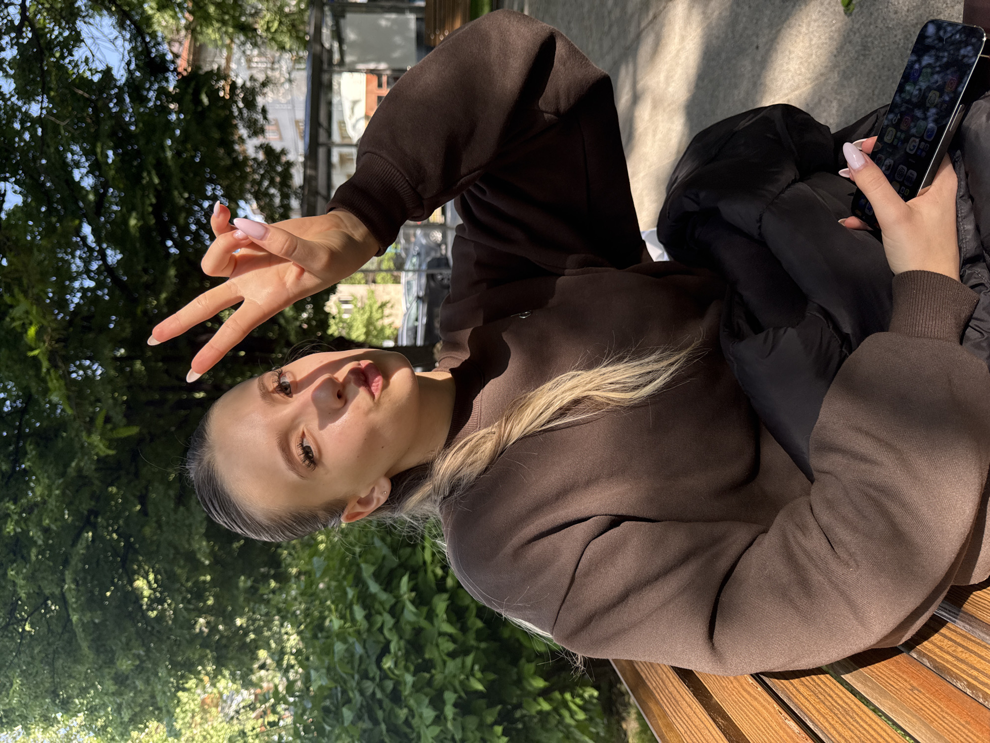A young woman is seated on a wooden bench in Plovdiv, Bulgaria, holding a black mobile phone in her right hand. She is wearing a brown sweatshirt and a black puffer jacket. She is holding up her left hand with her fingers making the peace sign. The background includes trees and foliage, indicating a park or green space. The photo is taken during daylight, with sunlight filtering through the trees.