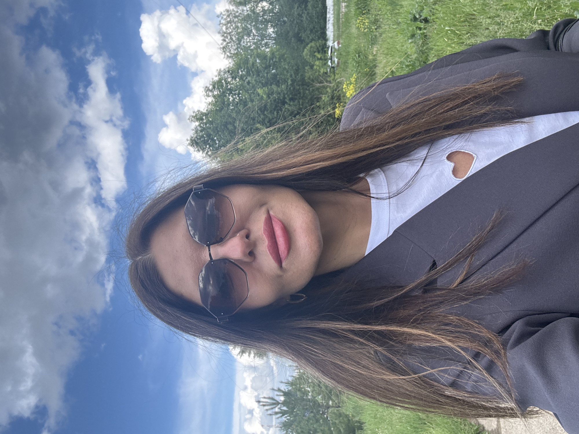 This close-up outdoor portrait, likely a selfie, features a young woman set against a natural backdrop in Chaikovsky, Russia. The image is oriented with the woman's head on the left and her body extending to the right.
The central subject is a woman with long, flowing dark brown hair. She is wearing dark, geometric-framed sunglasses and has pink lipstick applied to her lips, with a subtle, serene expression and a hint of a smile. Her attire consists of a white t-shirt featuring a heart-shaped cut-out on the chest, layered under a dark grey or black blazer. A small gold-colored earring is partially visible on her right ear.
The background suggests a clear, pleasant day outdoors. On the left side of the frame, a vibrant blue sky with scattered white clouds dominates. To the right, a lush green landscape unfolds, characterized by trees and grass, with hints of yellow wildflowers. In the distance, beyond the trees, there appears to be a body of water, possibly a river or lake, with a light-colored structure visible on its far bank. The scene captures a tranquil moment in a natural setting. No text is visible in the image.