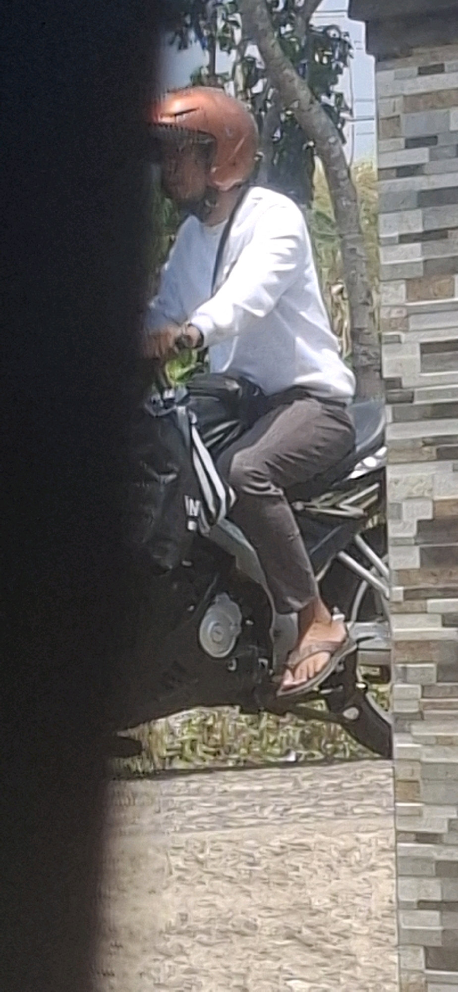 A person wearing an orange helmet and a white long-sleeved shirt rides a motorcycle. The person is wearing dark pants and light-colored sandals. A black bag with white stripes is attached to the back of the motorcycle. The motorcycle is parked on a gravel surface next to a brick wall. Trees and foliage are visible in the background. The image is blurry and taken from an oblique angle, with a dark, out-of-focus area obscuring the left side of the frame. The location is identified as Cibinong Hilir, Indonesia.