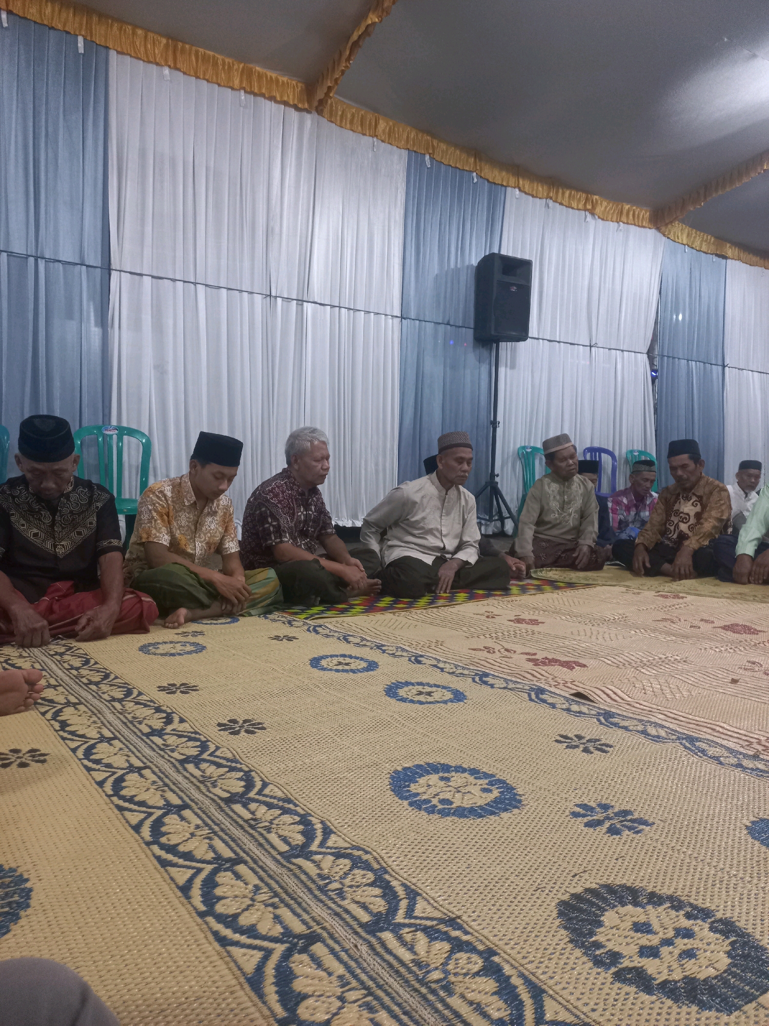 The image depicts a gathering of approximately eight men sitting on woven mats on the floor, inside a tent or covered venue. The men are dressed in traditional Islamic attire, including peci (caps) and long-sleeved shirts or robes, some with sarongs. They are seated in a relaxed, informal manner, with most facing towards the left of the frame. A large speaker is visible mounted on a stand in the background, behind a backdrop of blue and white curtains. Green plastic chairs are also visible behind some of the men. The mats on the floor have a patterned design in blue and beige. The overall scene suggests a religious or community gathering taking place.