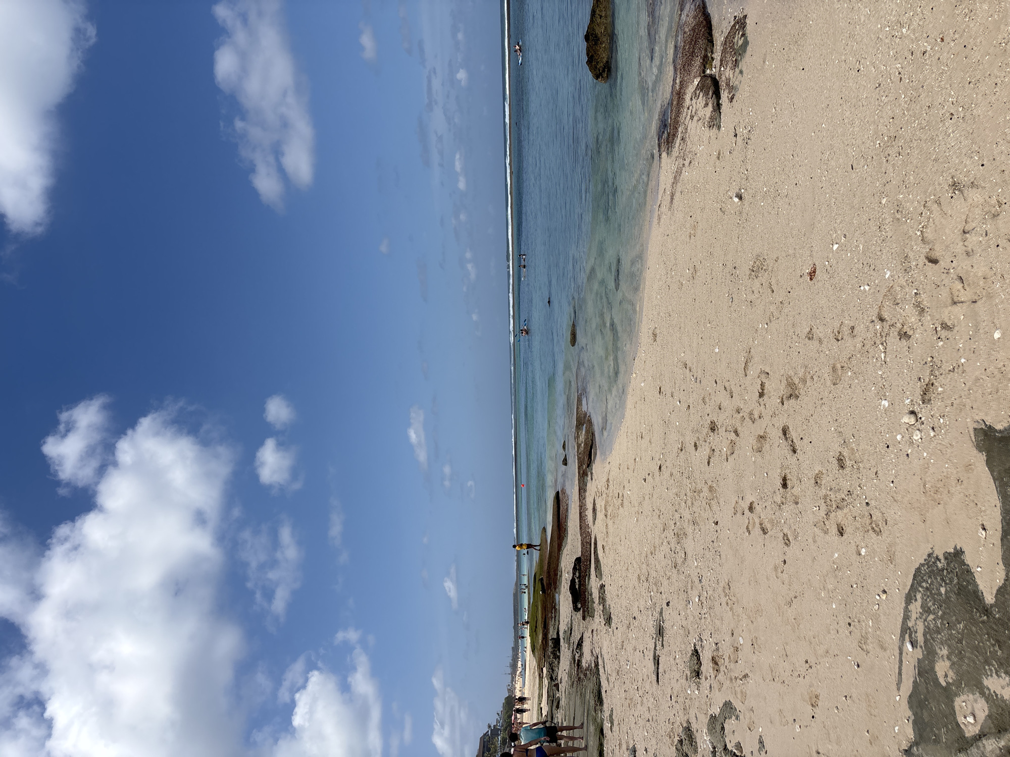 The image depicts a sun-drenched beach scene at an unspecified location in Kuta Selatan, Indonesia. The foreground is dominated by a sandy beach, dotted with small shells and pebbles, and imprinted with numerous footprints. Several dark, wet rocks are scattered across the sand and at the water's edge.
In the middle ground, the shallow, clear turquoise water laps gently at the shore, revealing submerged rocks and sand. A few people are visible in the water and on the beach. To the left, two individuals, one wearing a blue bikini and the other in dark shorts, walk along the water's edge. Further along the beach, a line of people are walking and standing, some near a resort-like structure visible in the distance. A lone figure in a yellow shirt stands on a large, dark, moss-covered rock in the middle of the water.
In the background, the ocean stretches out to the horizon, marked by a line of breaking waves. Several individuals are engaged in water activities, including what appear to be people on surfboards or paddleboards further out in the ocean. The sky above is a vibrant blue, populated by scattered white cumulus clouds. The overall impression is of a typical, bustling tropical beach environment.