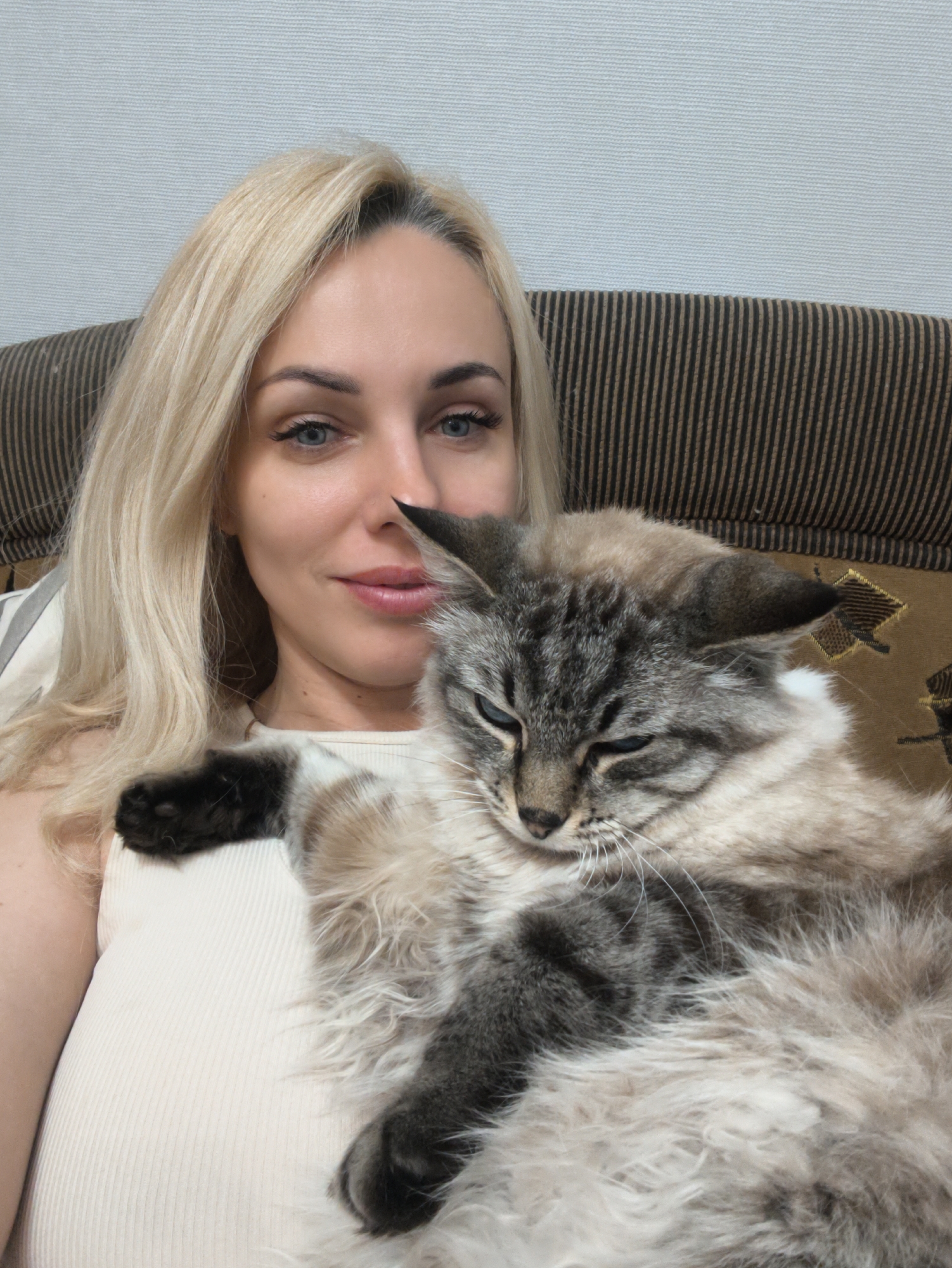 A woman with blonde hair is holding a cat. The woman is fair-skinned with blue eyes. She is wearing a light-colored tank top. The cat is a long-haired breed with gray and beige fur. The cat has blue eyes and is resting its paw on the woman's chest. They are sitting on a striped couch. The background is a plain wall. There is no evidence of being in the city of Sevastopol, Ukraine.