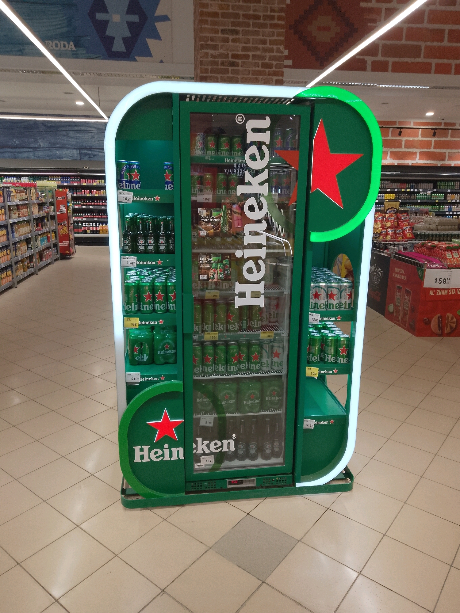 This image depicts a retail store, likely a supermarket or hypermarket, in Belgrade, Serbia. The main subject is a prominent green Heineken beer cooler, which is brightly lit from within and has a large Heineken star logo on its side. The cooler is filled with rows of green Heineken beer cans and bottles. Shelves stocked with various products are visible in the background, extending down a long aisle. The flooring is tiled in a beige and off-white pattern. The store appears to be well-lit, with overhead lighting illuminating the space. The overall impression is that of a modern retail environment.