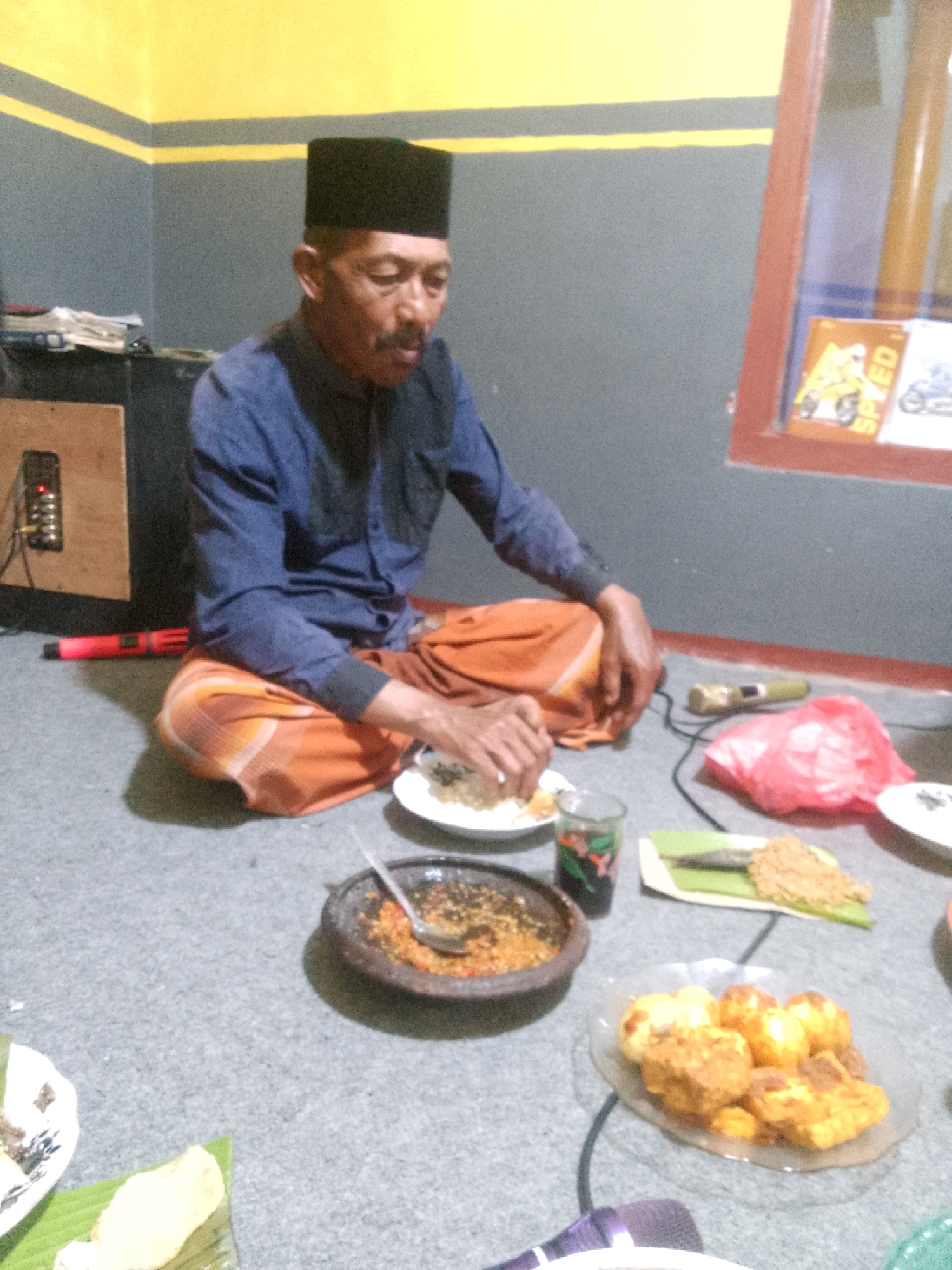 The image depicts an elderly man wearing a blue shirt and an orange sarong, seated on the floor in what appears to be a room in Krucil, Indonesia. He is looking down at a plate of food in front of him. Several other plates of food, a glass with a dark liquid, a red plastic bag, and a mortar with a pestle containing chili paste are also present on the floor, suggesting a meal is being shared or prepared. A speaker, a red object that could be a stick or tool, and a microphone are visible in the background, as well as a window with some decorative items. The walls are painted grey with yellow stripes. The man's posture and the surrounding food indicate a casual, possibly communal dining setting.