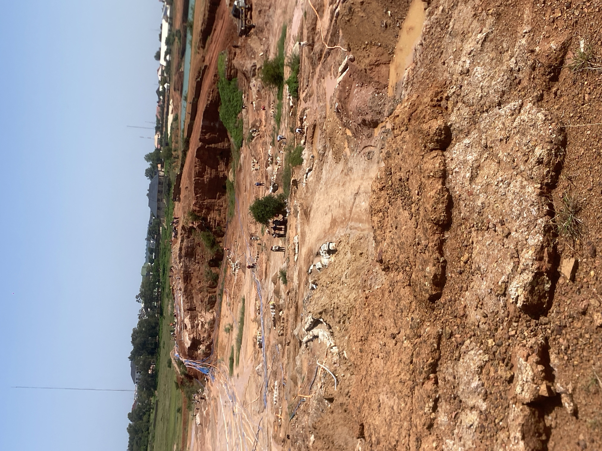 The media file depicts an aerial view of a mining or excavation site in Jos, Nigeria. The scene is characterized by reddish-brown earth, likely a quarry or a large construction site, with several people scattered throughout. The terrain is uneven, with steep inclines and muddy areas, suggesting recent excavation.
Multiple individuals are visible, seemingly engaged in labor. They are dressed in casual clothing, and some appear to be working with tools or carrying materials. The presence of various pipes, some blue and white, running across the site indicates some form of water or material transport system, possibly for the mining operation or construction. A truck is also visible, further suggesting industrial activity.
The setting appears to be outdoors during daylight hours, with a clear blue sky. The overall impression is one of active work and a large-scale operation. The landscape includes some vegetation, with trees and green grassy areas bordering the excavated zone, and buildings in the distance, indicating proximity to a populated area.
There is no visible text within the media file. The activity appears to be focused on extraction or construction, with the raw earth and the presence of workers and equipment being the central elements. The atmosphere conveyed is one of hard work under natural conditions.
