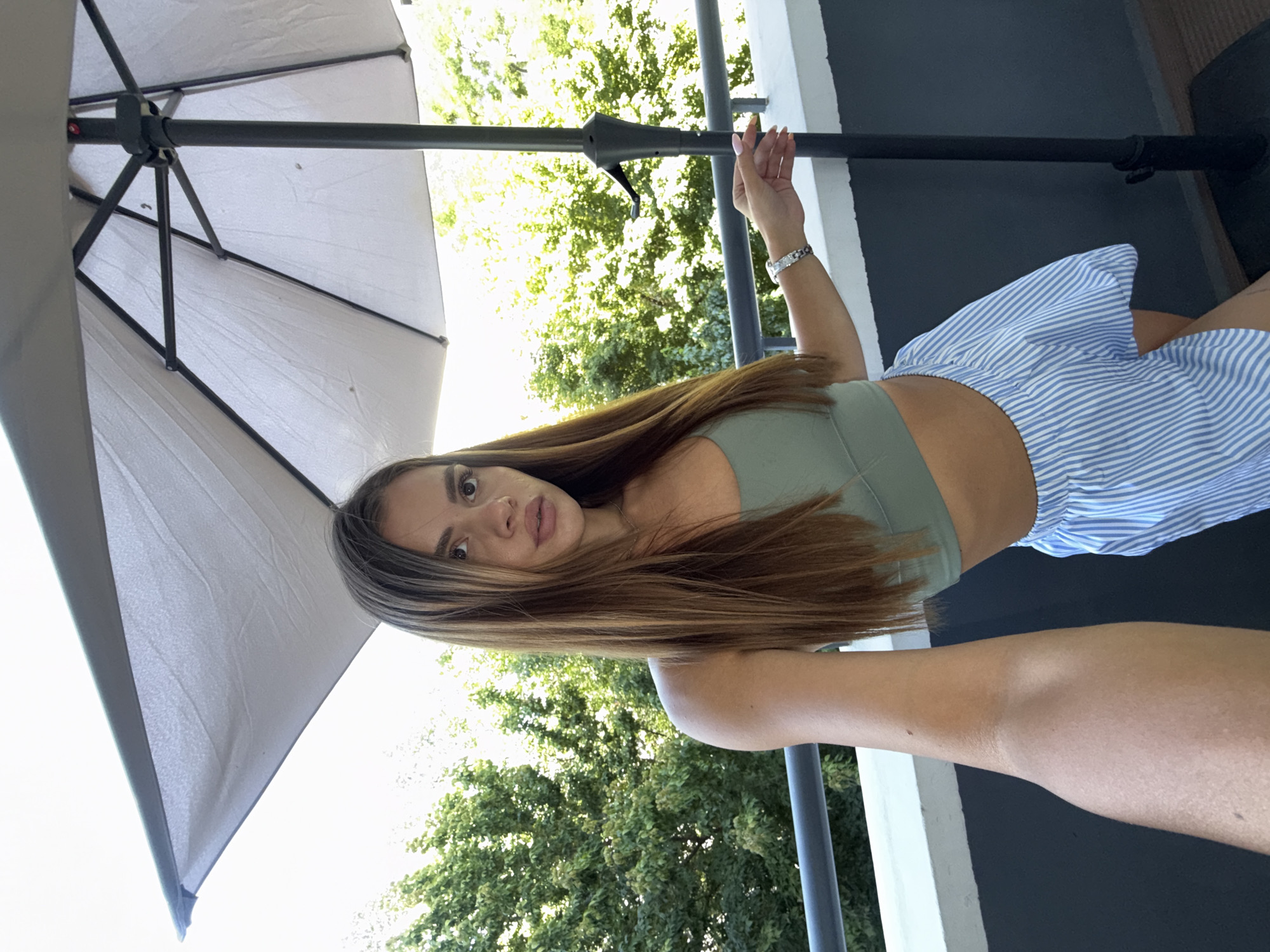 A young woman with long brown hair, wearing a green crop top and blue and white striped shorts, poses for a selfie on a balcony. She is holding her phone with her left arm, extending it towards herself to capture the shot. Her right hand rests on the pole of a large, beige outdoor umbrella. Behind her, lush green trees and foliage are visible, suggesting a location with ample greenery. The balcony railing is visible in the foreground, along with a dark gray wall. The scene appears to be outdoors, possibly in a residential area or a hotel, given the balcony and umbrella. The lighting indicates it is daytime, with bright natural light. The overall setting is casual and relaxed, suggesting a leisure activity or a moment of enjoyment on a warm day.
