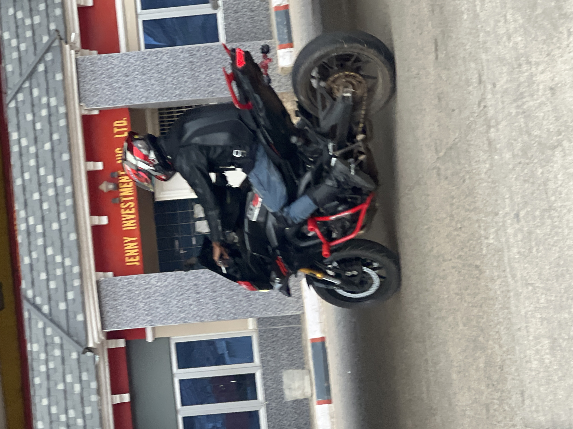 The image displays a person standing next to a black sport motorcycle, parked alongside a building. The person is wearing a black motorcycle jacket, blue jeans, and a distinctive red, black, and white full-face helmet. They are leaning over the motorcycle, seemingly making an adjustment or preparing for a ride.
The motorcycle is predominantly black with bright red highlights, possibly indicating a crash frame or specific styling elements. A gold-colored chain is visible on the rear wheel's sprocket.
The scene is set outdoors, on what appears to be a paved surface, likely a street or parking area. Adjacent to the motorcycle is a multi-story building with a red lower section and a gray textured upper facade. Several windows with blue-tinted glass are visible.
A prominent vertical sign on the red part of the building clearly displays the text "JENNY INVESTMENT LTD."
The photograph appears to have been taken during daylight hours under clear weather conditions, as indicated by the natural light and lack of visible precipitation. No specific time of day is discernible. The person's face is obscured by the helmet, so no emotions are evident. The overall activity suggests a brief stop or pre-ride check. The location context is Gura Top, Nigeria.