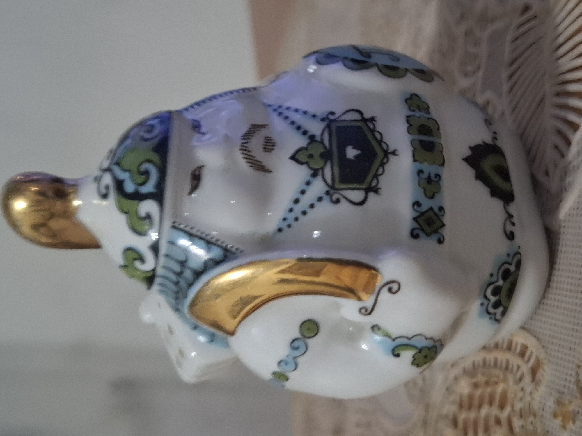 A close-up, side-view, eye-level shot shows a ceramic figurine of a rotund man with a golden headpiece. The figurine is white with blue and green decorative patterns, including a shield-like emblem on the chest and floral motifs along the base. The man has a painted mustache and closed eyes. The figurine is placed on a textured, light-colored surface, possibly a tablecloth or doily, with a blurred background of a light-colored wall. The lighting is soft and diffused, casting minimal shadows. No specific location in Almaty, Kazakhstan, can be identified from the image.