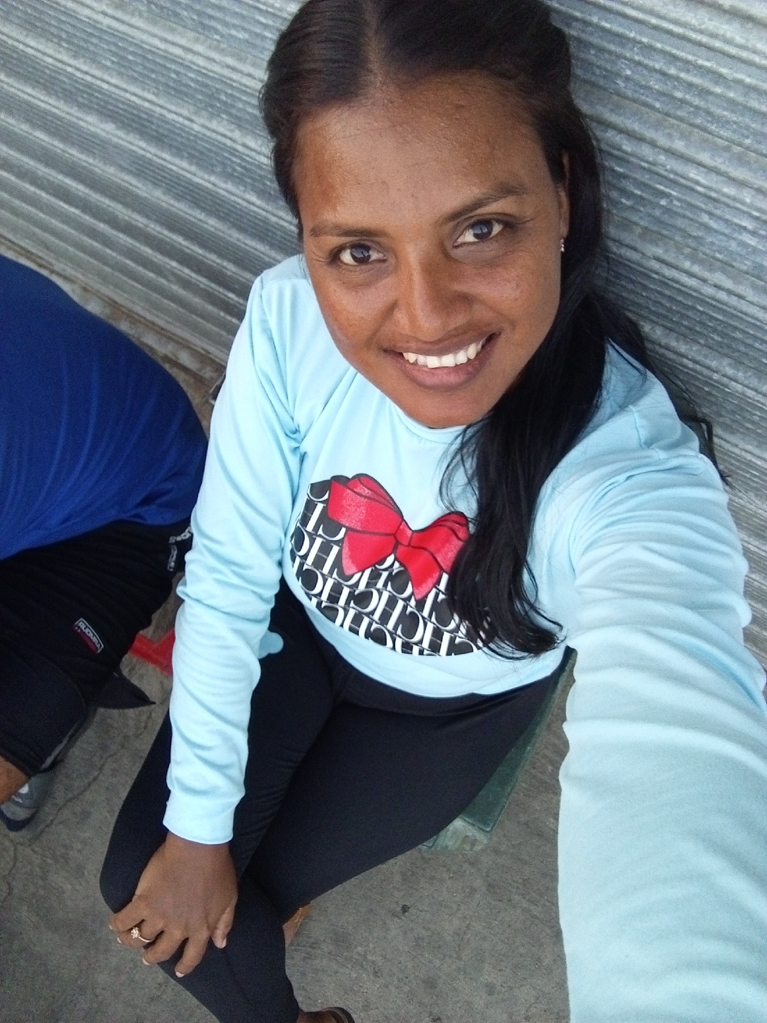 A woman with dark hair is smiling at the camera while sitting in front of a wall. She is wearing a light blue long-sleeved shirt with a red bow design, and black leggings. A man is seated beside her, wearing a blue shirt and black shorts. The scene is located in the city of Unknown, Unknown.