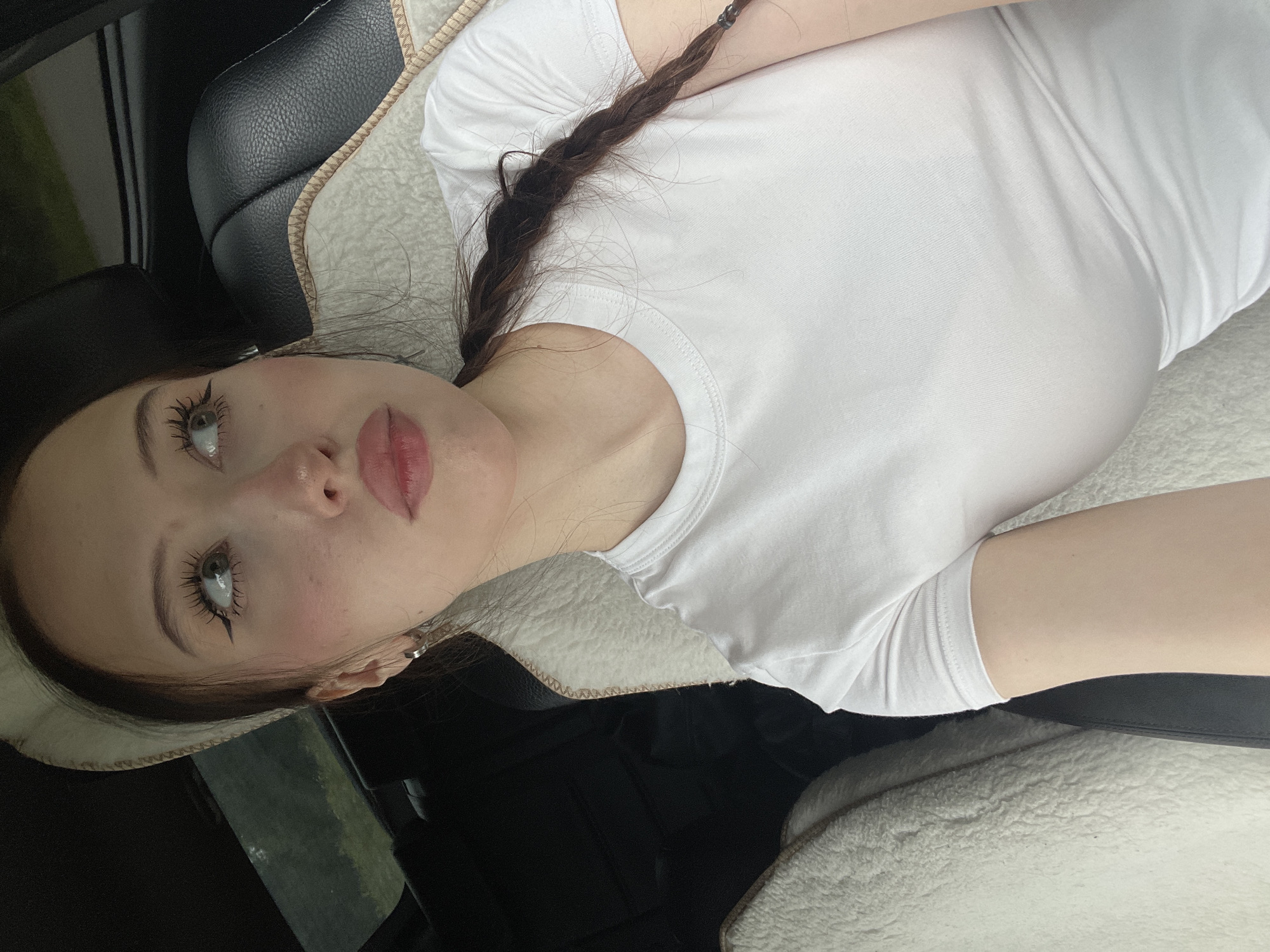 A young woman with light skin, long dark brown hair, and green eyes is sitting in the passenger seat of a car. She is wearing a white t-shirt and a light-colored headband. Her makeup includes winged eyeliner and pink blush. Her hair is in a braid that hangs over her shoulder. The car's interior is visible, including a black leather seat with a white fuzzy liner. The scene is not in Gavrilovka, Russia.